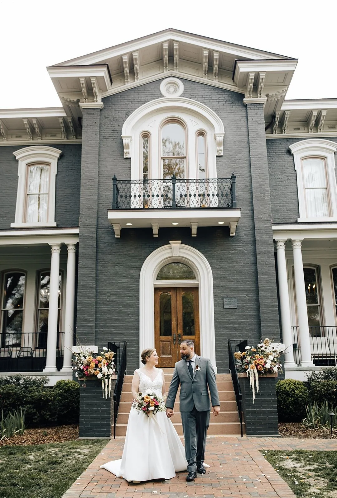 Couple walking hand in hand in front of Heights House in Raleigh, framed by wedding florals on the historic front steps