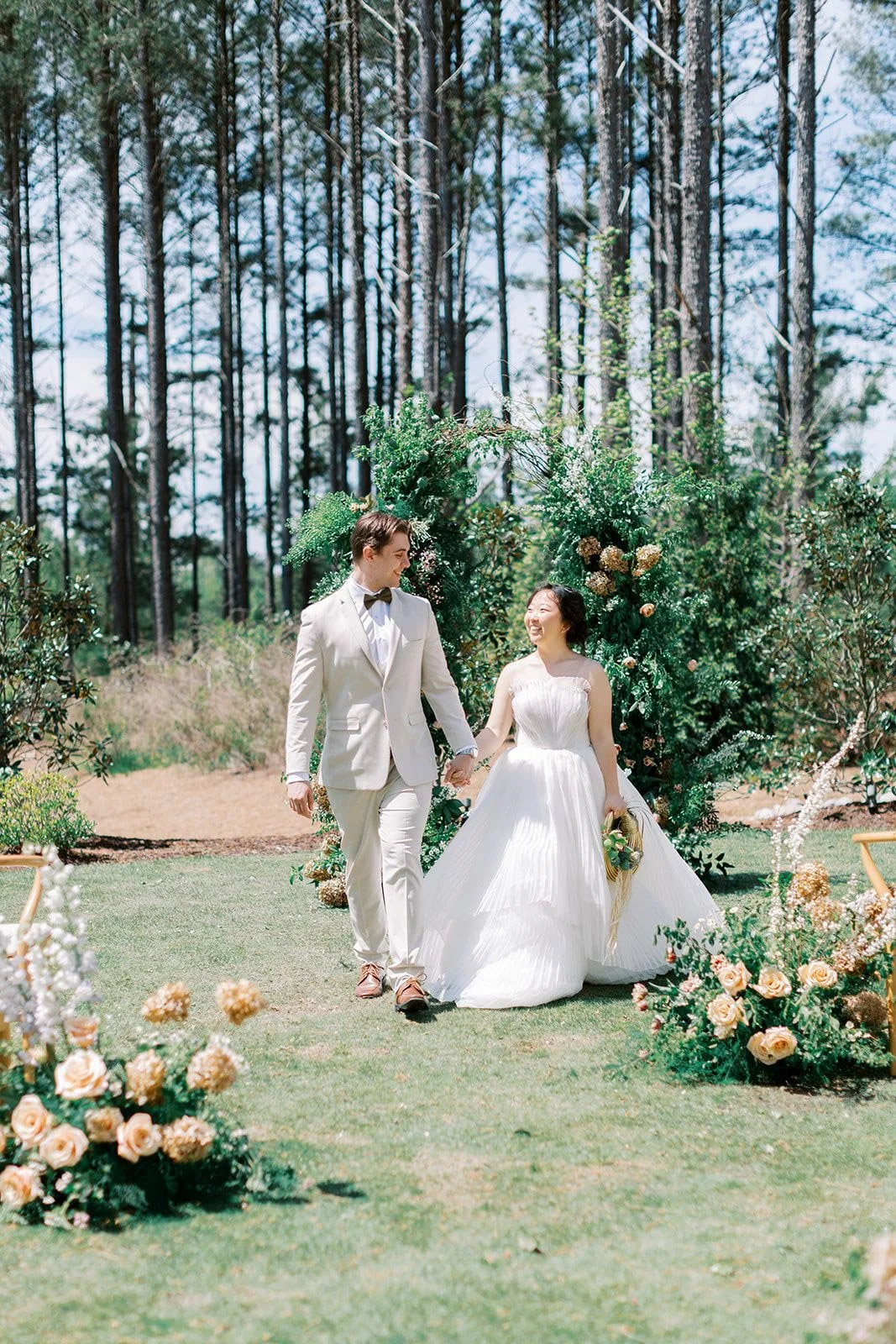 A couple walks hand in hand down the aisle beneath a lush greenery ceremony installation designed by Wild Flora Flowers at The Upchurch wedding venue in Raleigh, North Carolina.