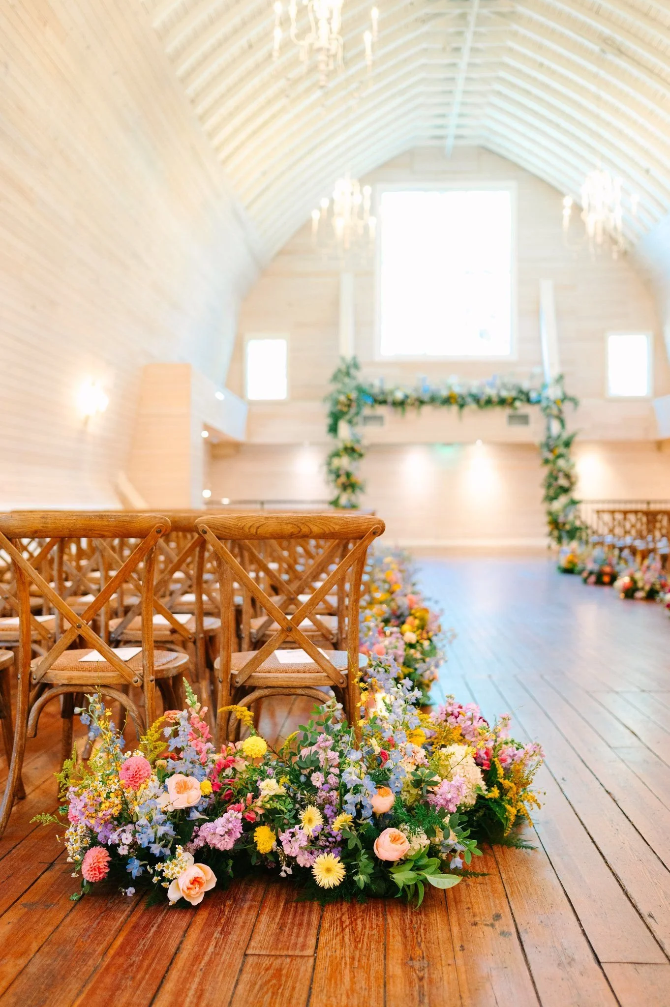 Colorful flowers lining a wedding aisle, leading to a larger than life vibrant flower arch inside of a historic barn venue