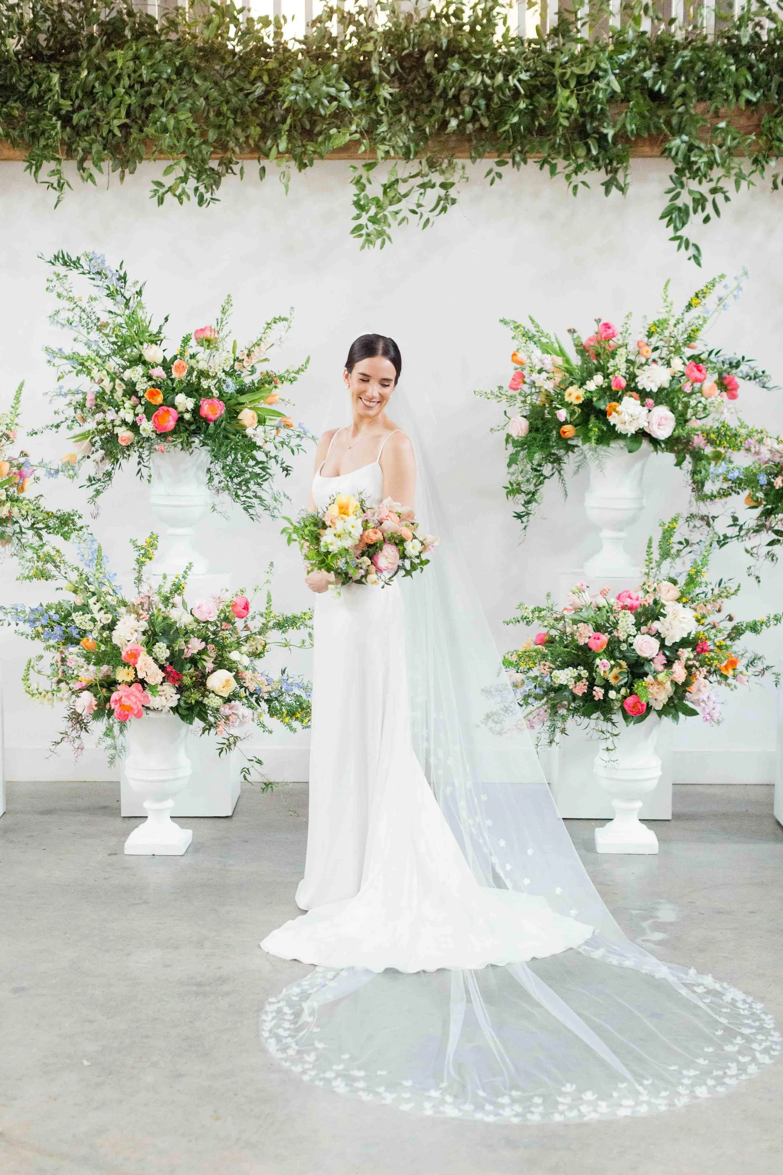 Bride in a minimalist wedding gown holding a bouquet, standing in front of a floral installation with urn arrangements at The Barn of Chapel Hill