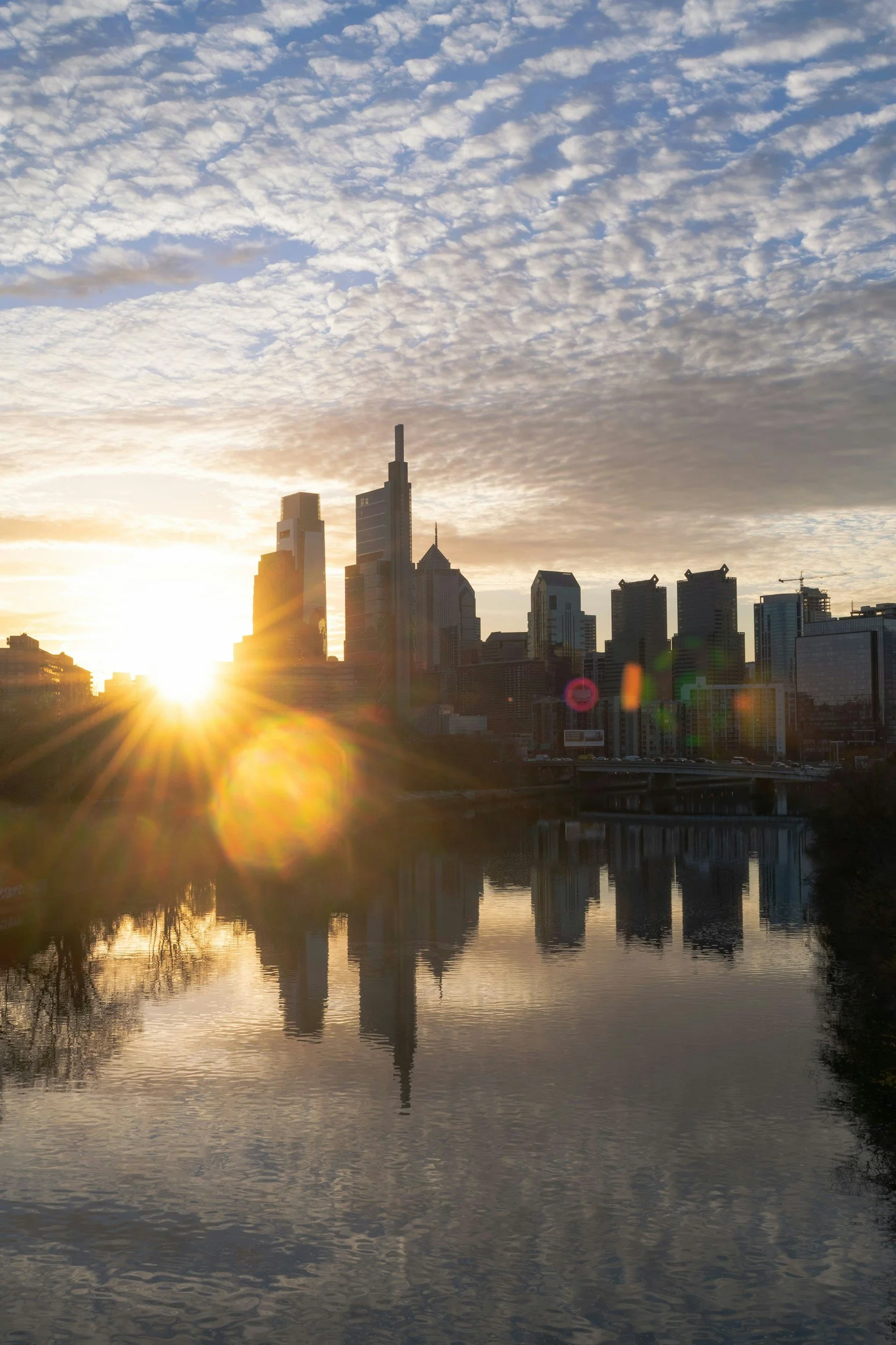 Sunset over a city skyline with tall buildings and a river reflecting the buildings and sky.
