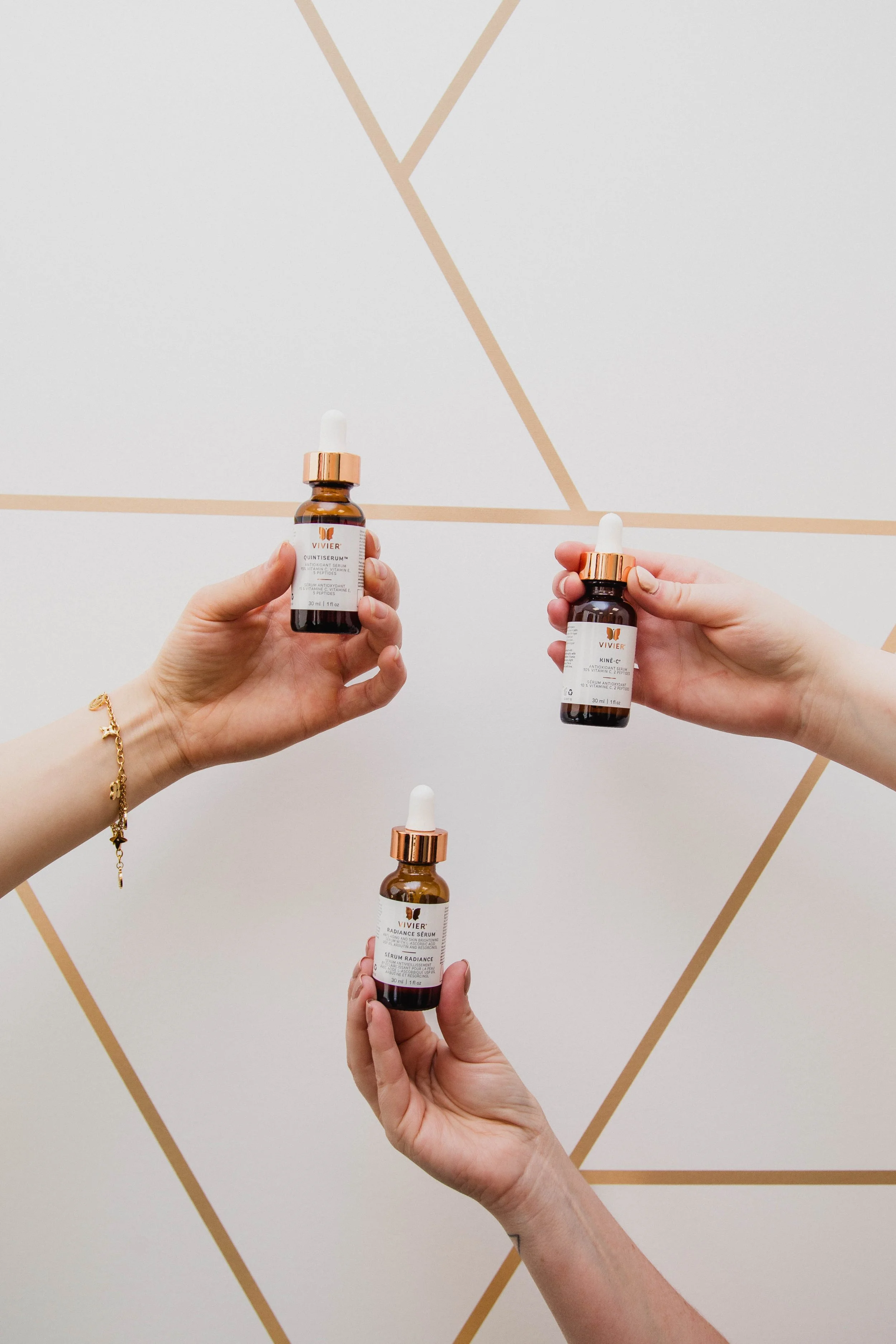 Three hands holding skincare serum bottles against a geometric patterned wall. The bottles are brown with white labels and dropper tops.