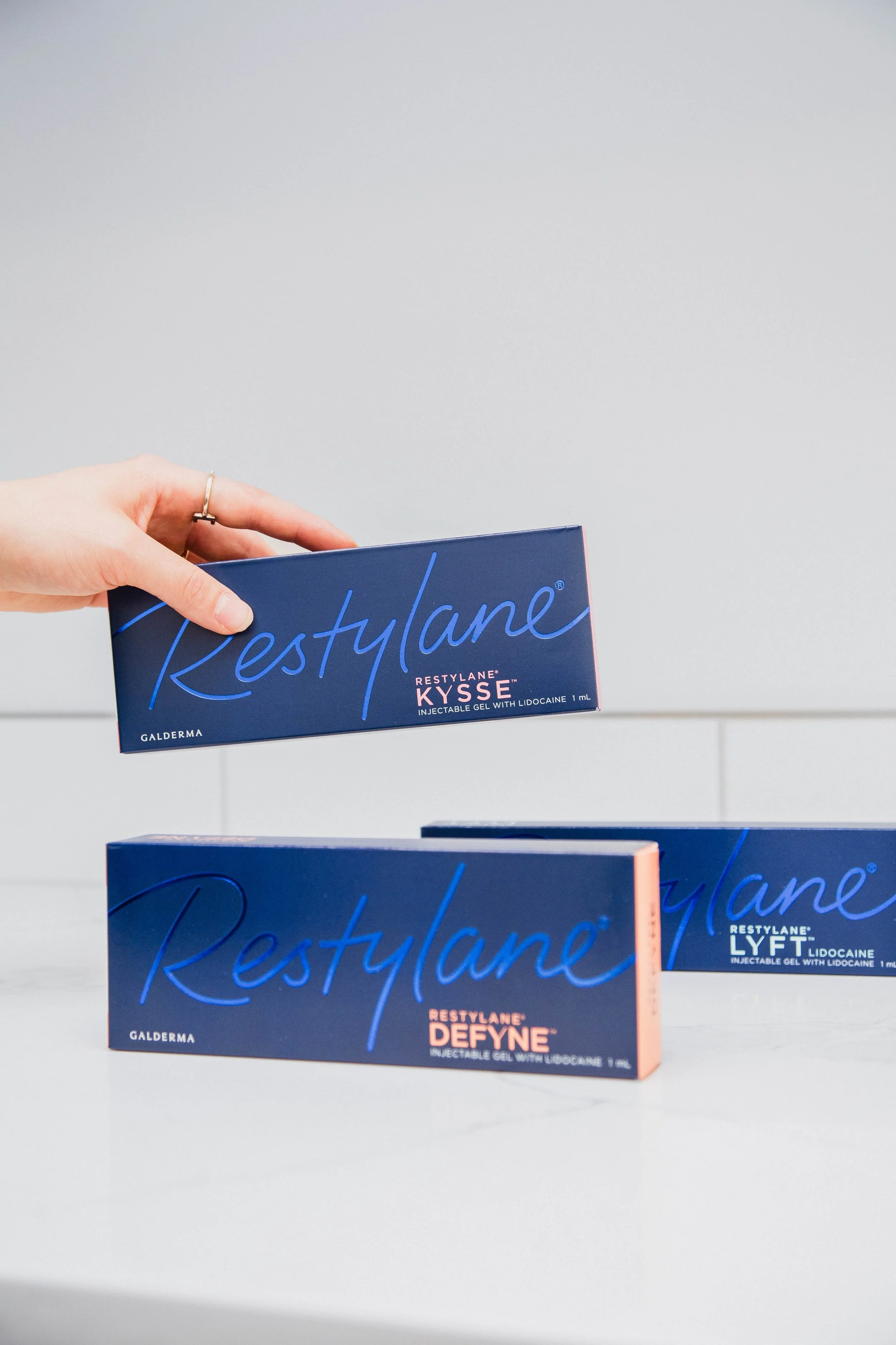 A hand holding a box of Restylane KYSSe, with boxes of Restylane Defyne and Restylane Lyft on a white surface - HA fillers used to treat the signs of aging.