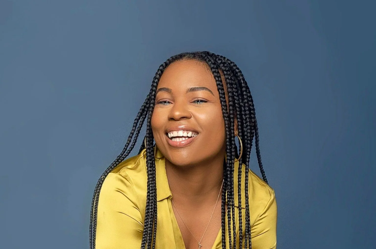 A woman with long black braids, gold hoop earrings, and a yellow satin shirt, smiling with a blue background.