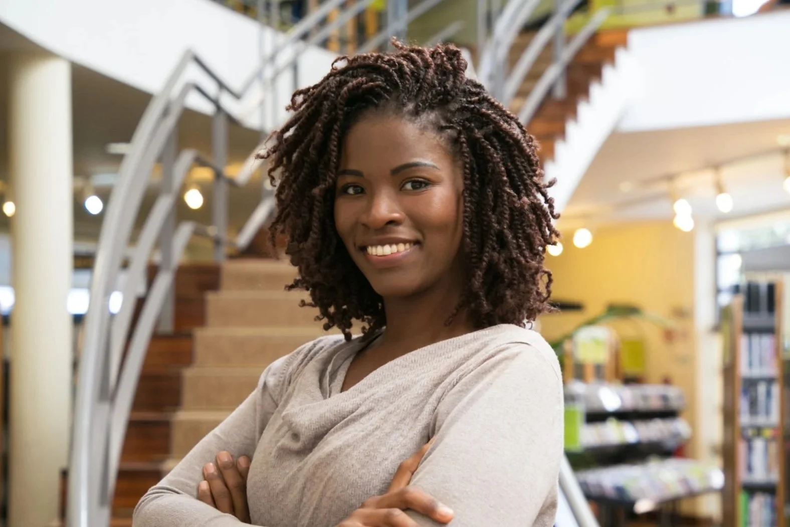 A woman with curly hair smiling, standing with arms crossed inside a library or bookstore.