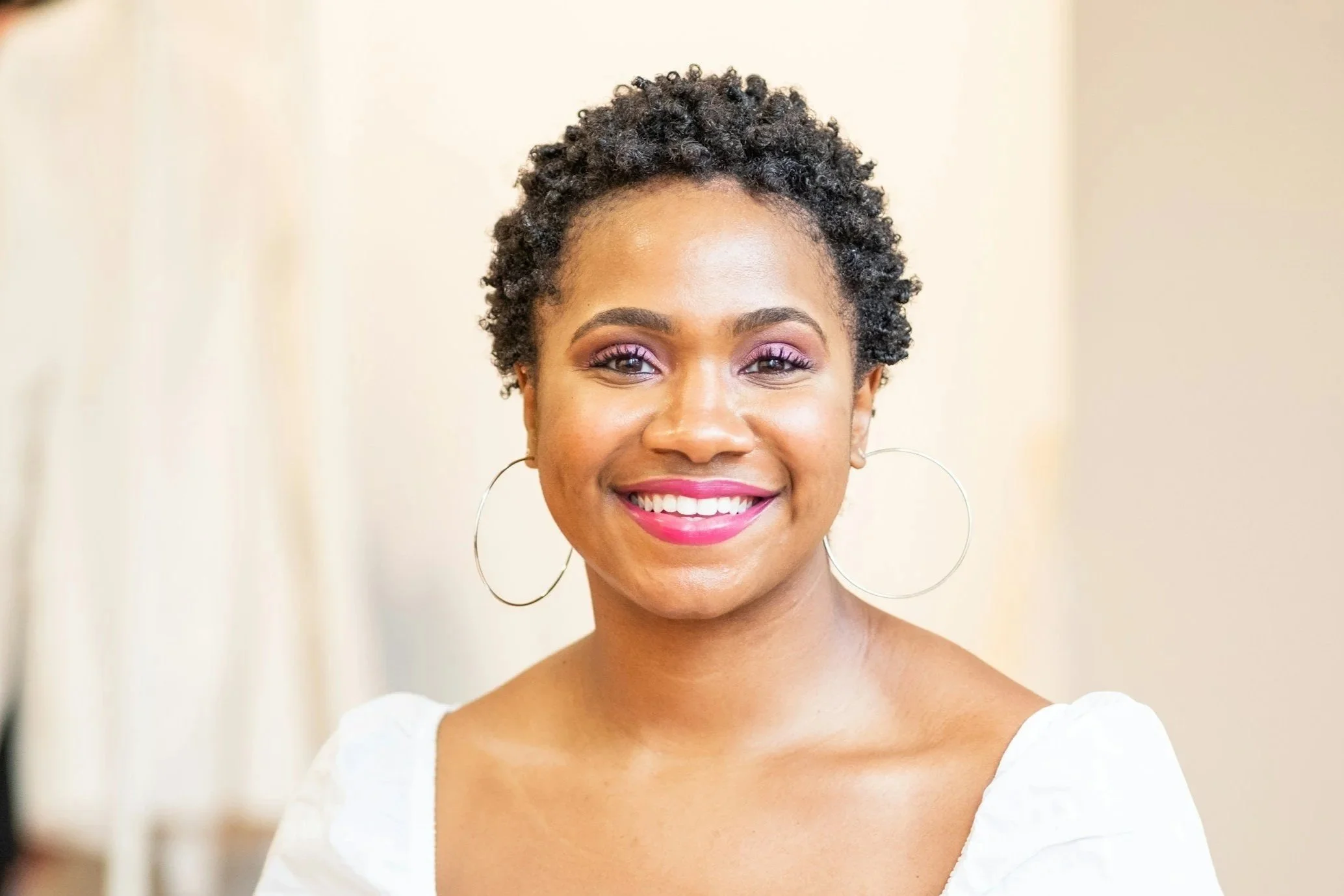 Close-up of a smiling woman with short curly hair, wearing hoop earrings and a white top, in a well-lit indoor setting.