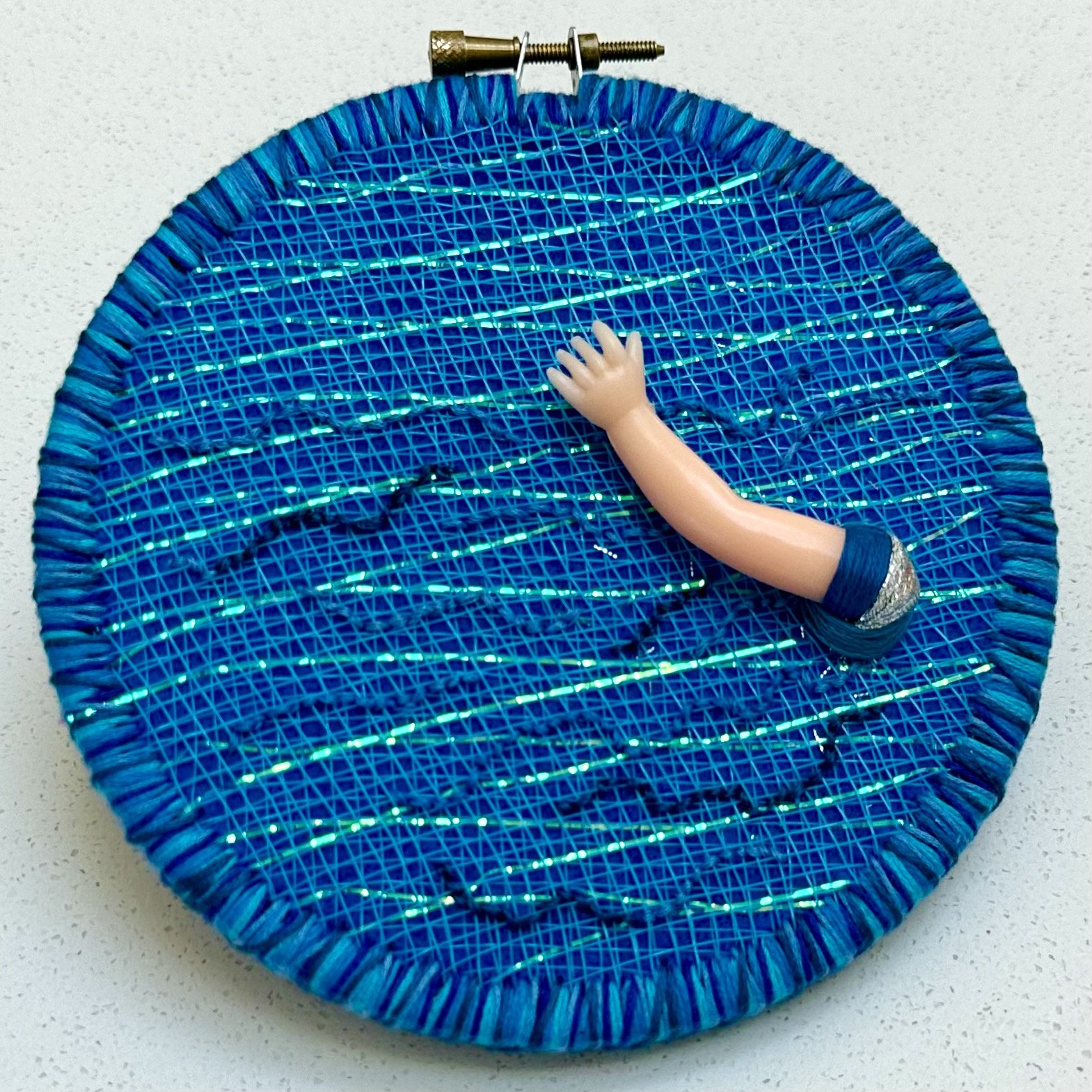 From Water: Meditation on Reaching Out - (2024) embroidery thread and plastic webbing on upcycled fabric and felt with plastic doll's arm 5.5" disc $
