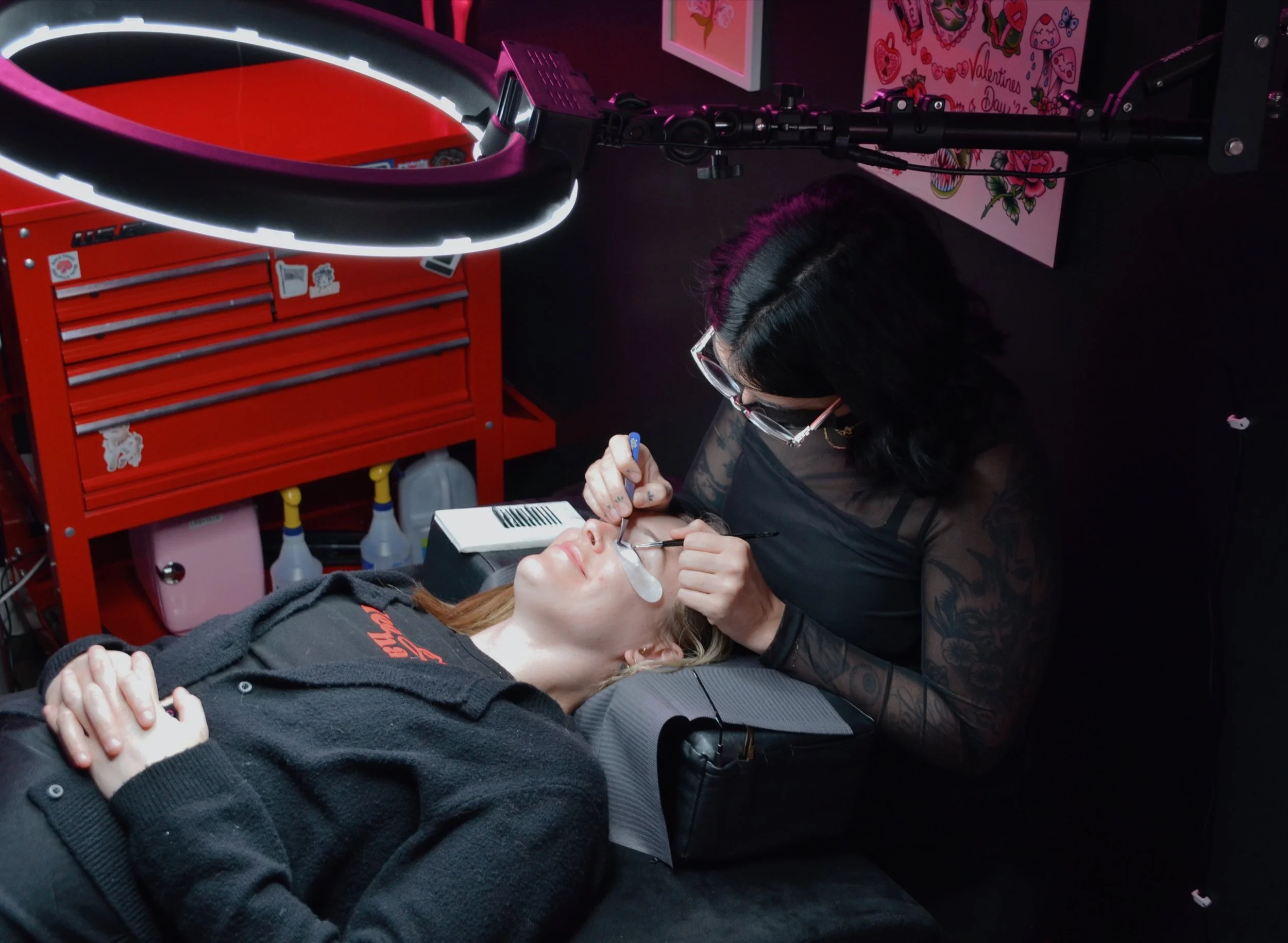 A woman with dark hair and glasses performing eyelash extensions on a client lying on a treatment bed in a tattooed black top salon. The client is wearing a black sweater with red text, and the woman is using tools under a magnifying lamp.