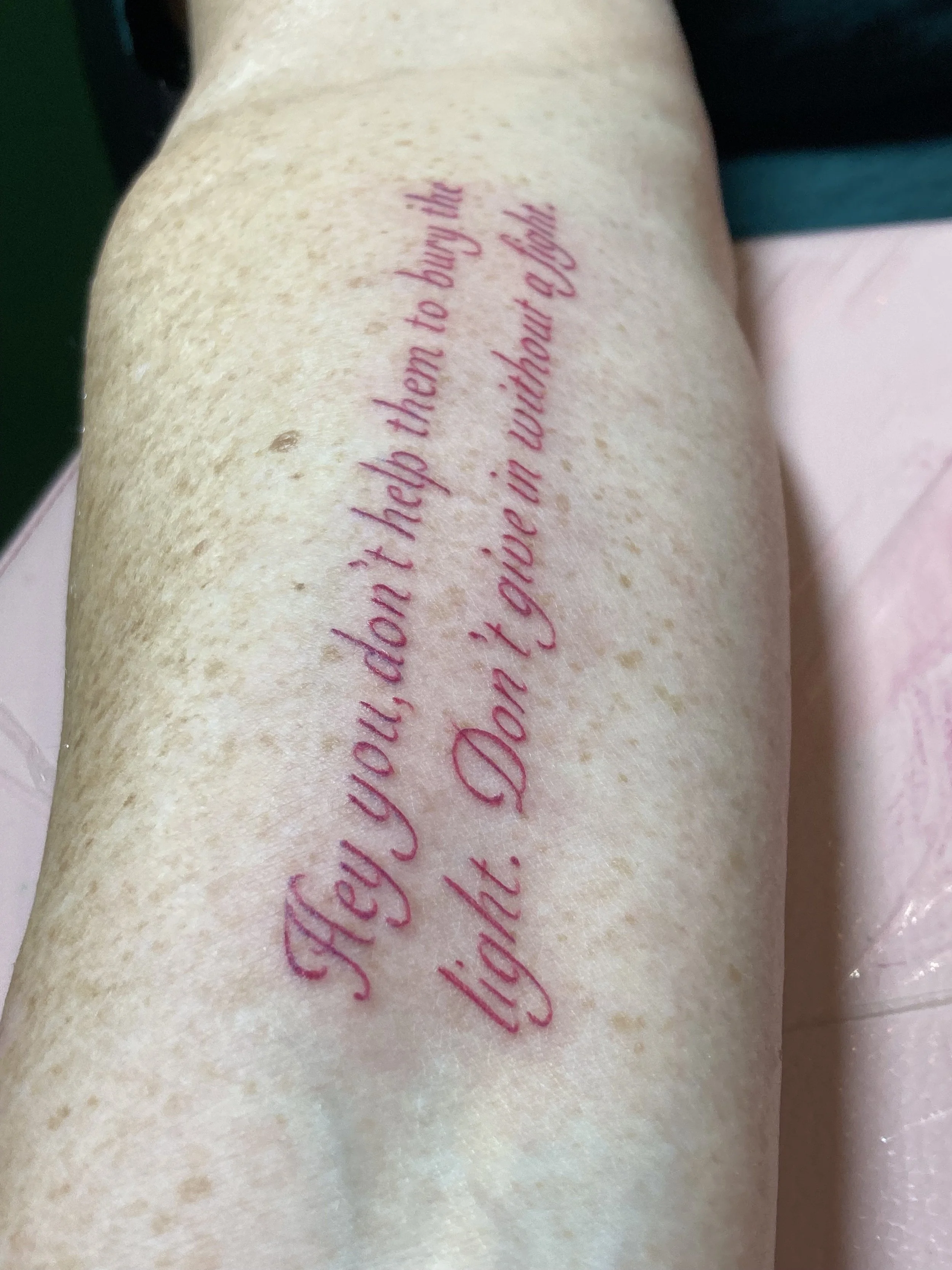 Tattoo on a person's leg with the quote, 'Hey you, don’t help them to bury the light. Don’t give up without a fight.' in pink cursive font.