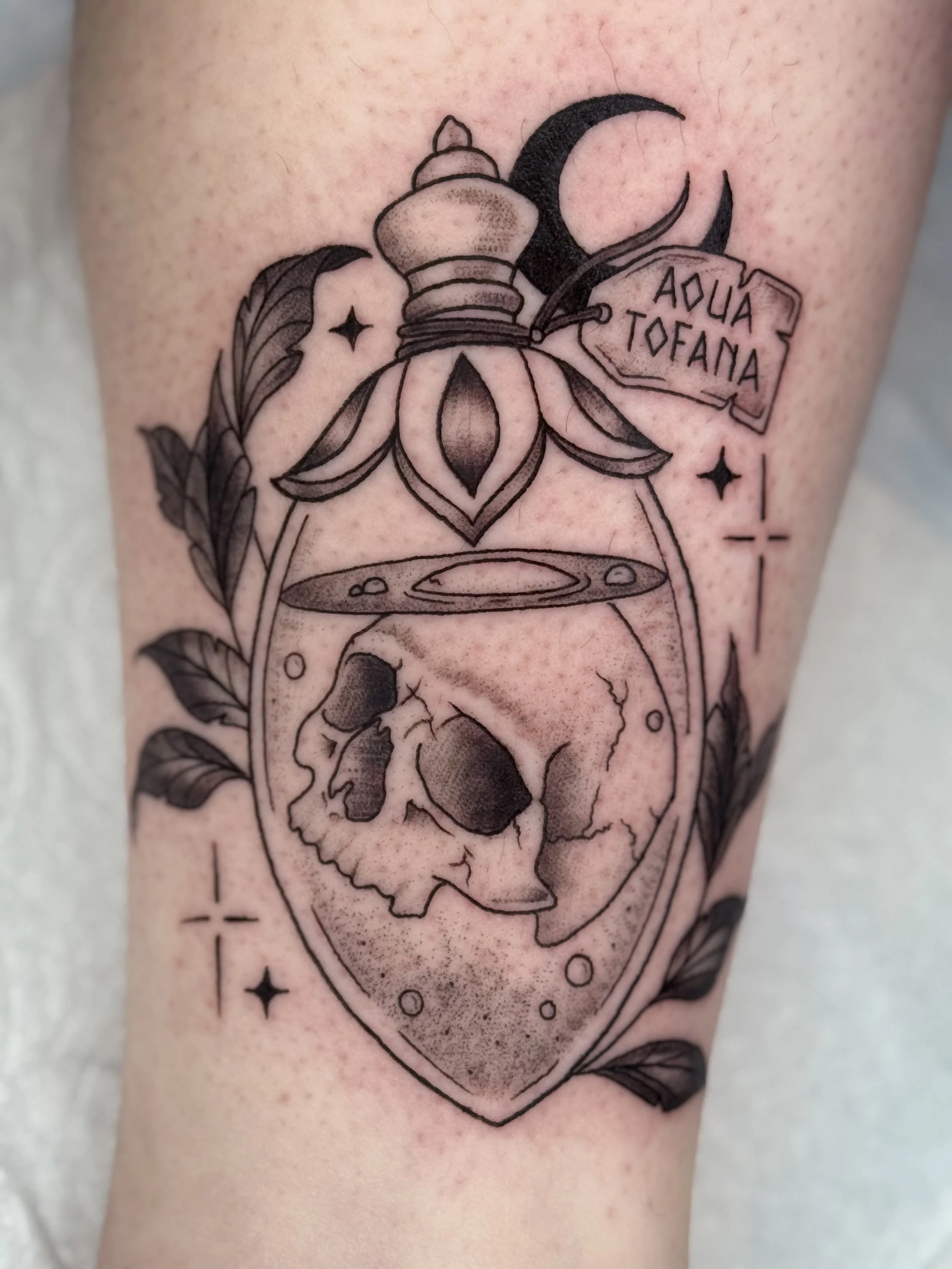 Tattoo of a perfume bottle with a skull inside it, surrounded by leaves, stars, and a sign that reads 'Aqua Toffania', all in black and gray ink.