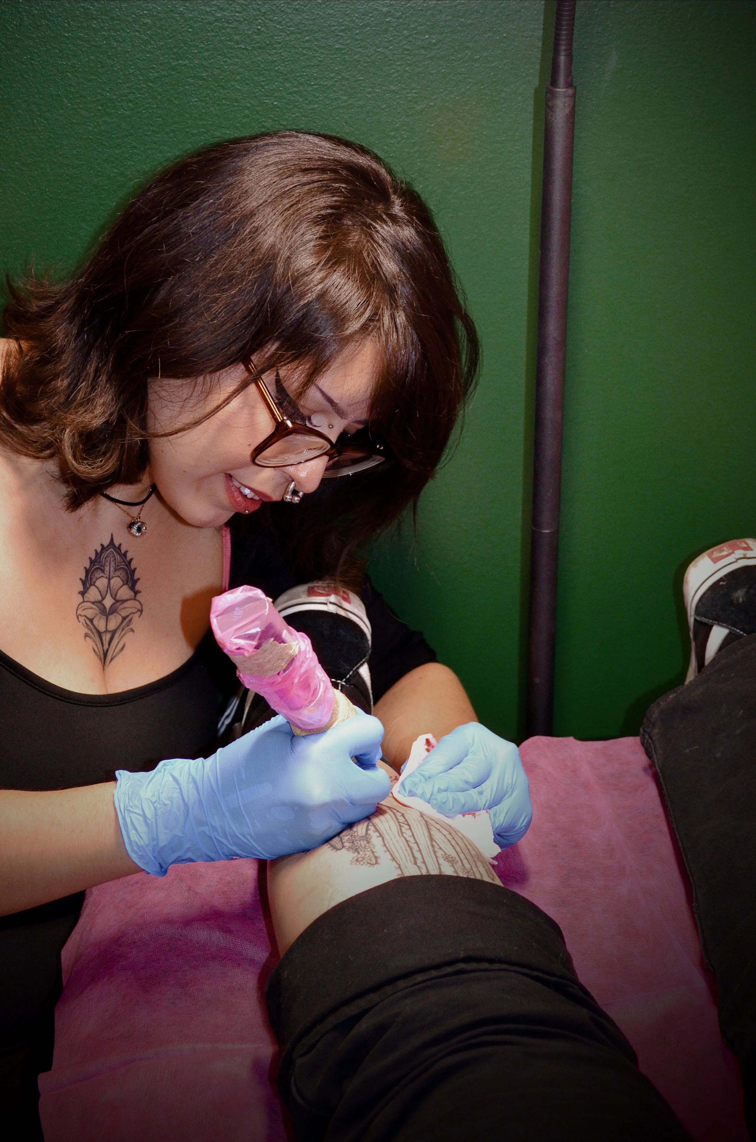 A tattoo artist with glasses and gloves is tattooing a design on a person's leg, who is lying down on a pink towel with a green wall in the background.