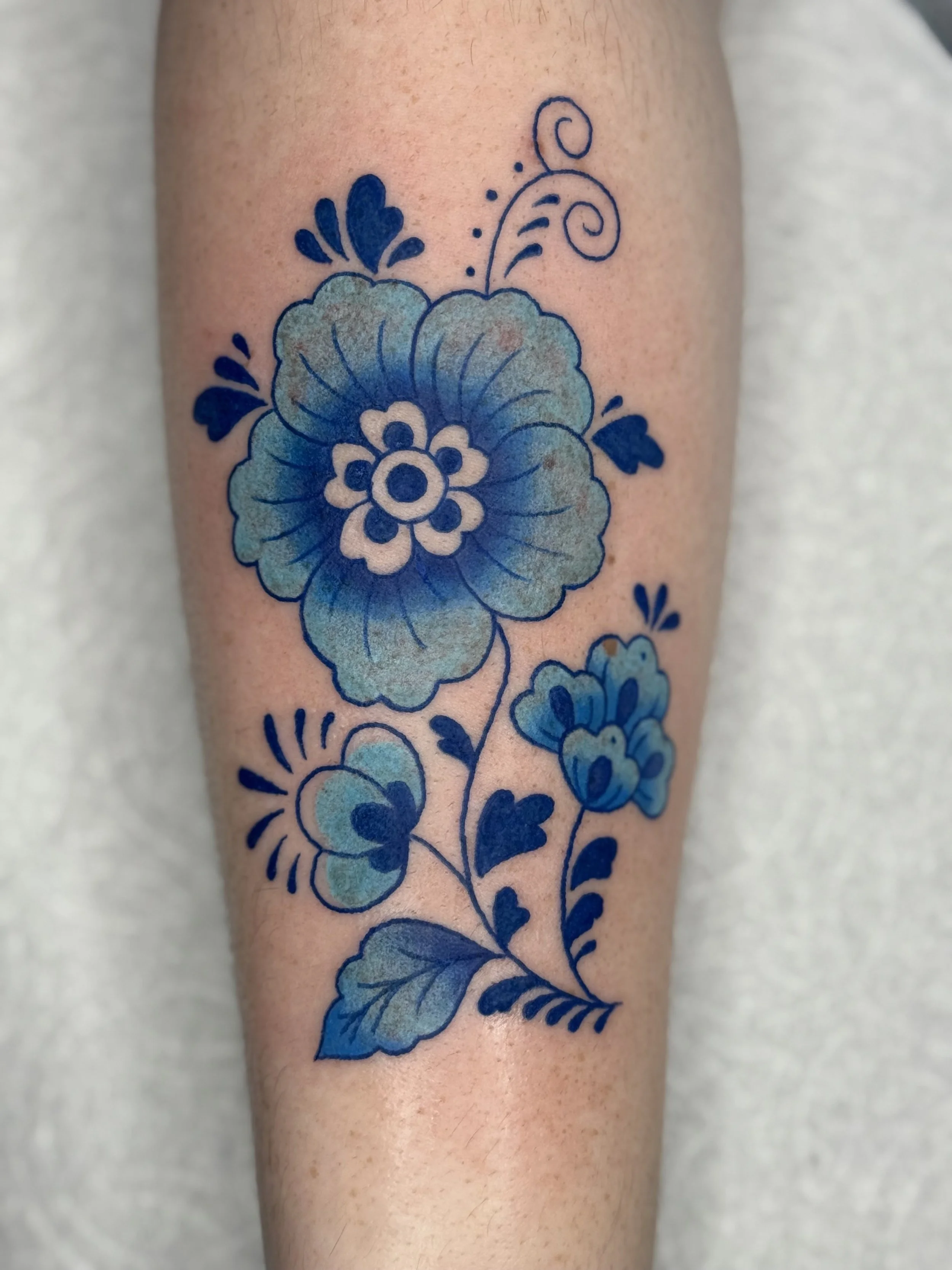 Blue floral tattoo featuring a large flower, smaller flowers, leaves, and decorative swirls on a person's arm.