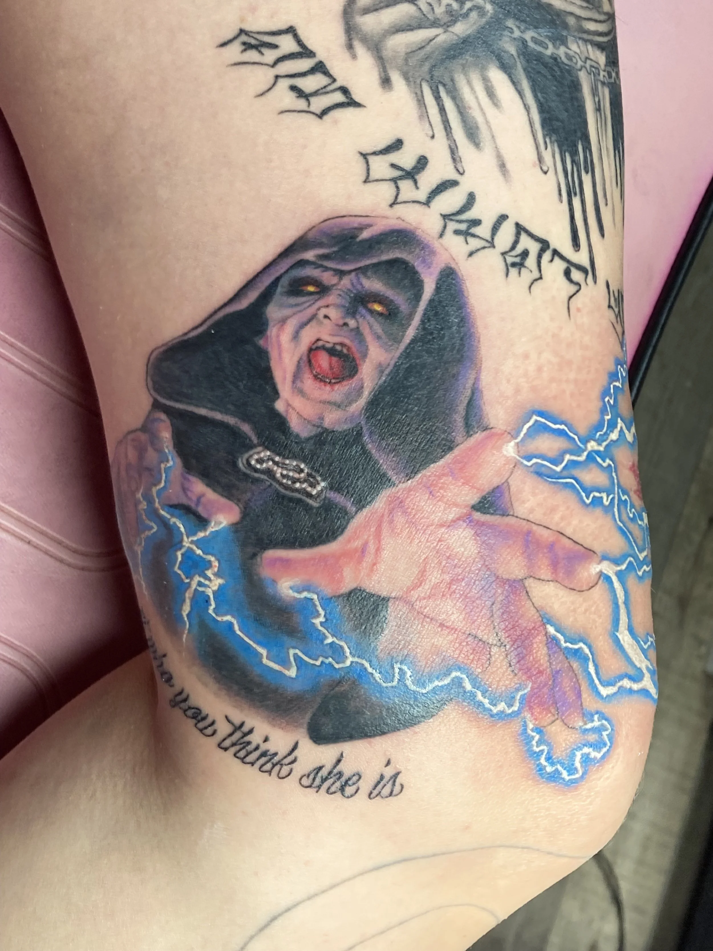 Tattoo of a witch-like figure with grey skin, glowing yellow eyes, and an open mouth showing fangs, reaching out with a hand surrounded by blue lightning. There is black Gothic-style text above and below the tattoo.