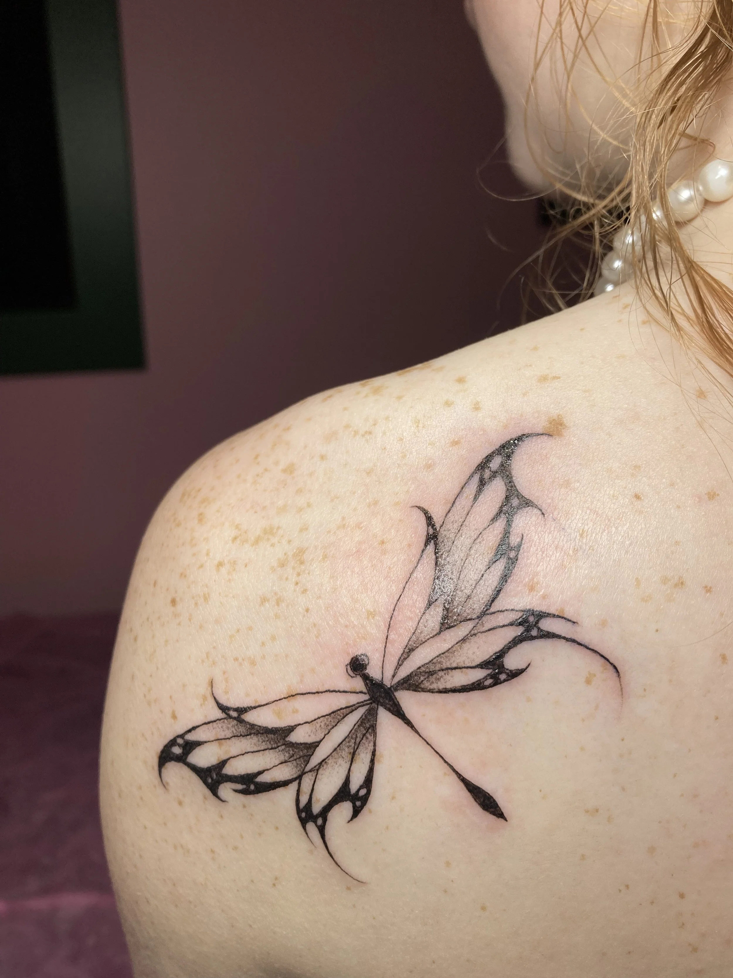 Tattoo of a butterfly on a person's shoulder with freckles and hair, wearing pearl earrings.
