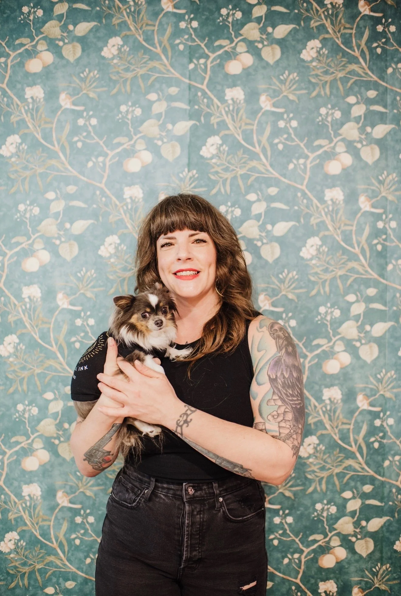 A woman with brown hair and tattoos holding a small black, white, and brown dog in front of a blue floral wallpaper background.