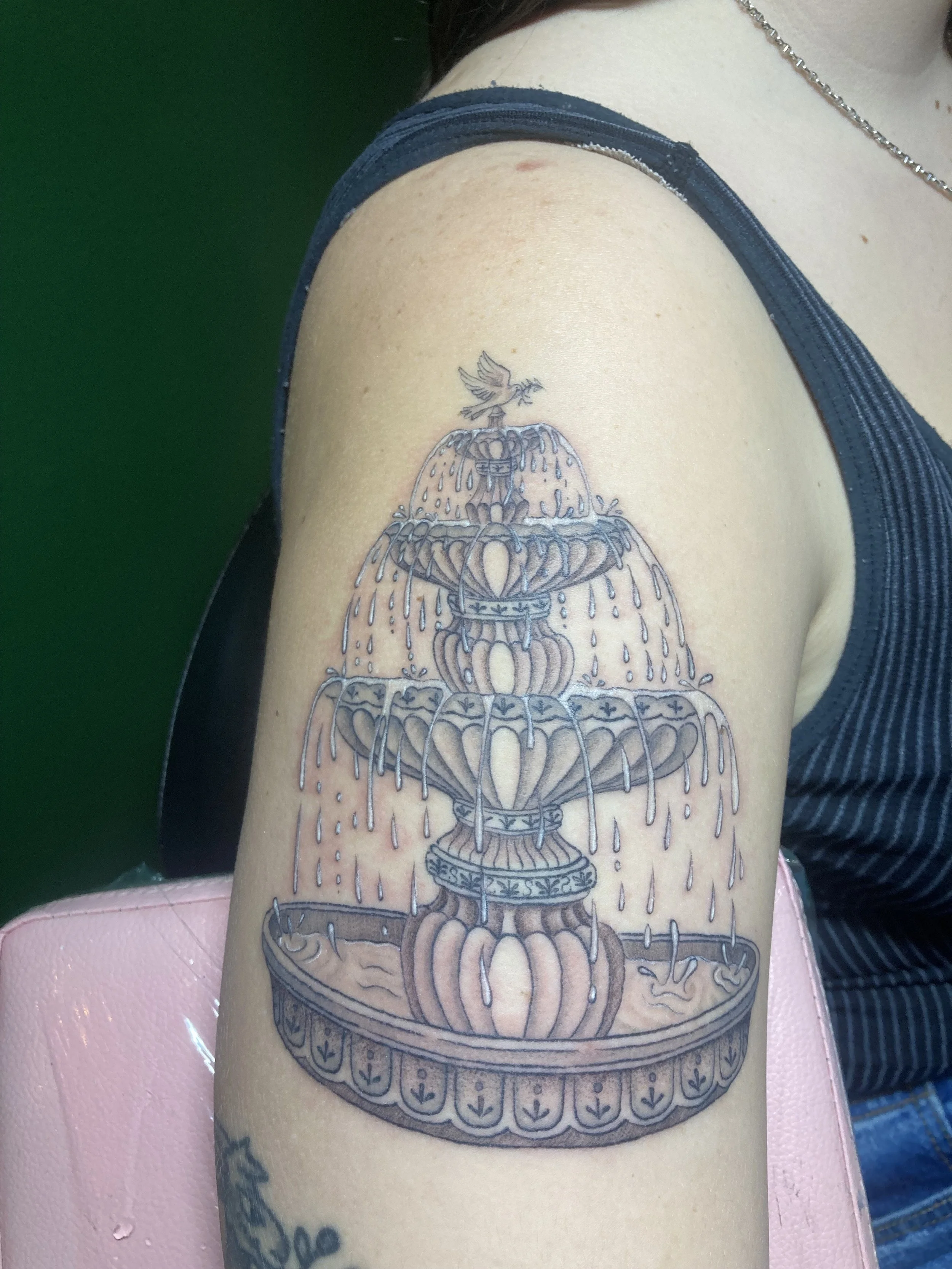 Tattoo of a water fountain with multiple tiers and a bird perched at the top, on a person's upper arm.