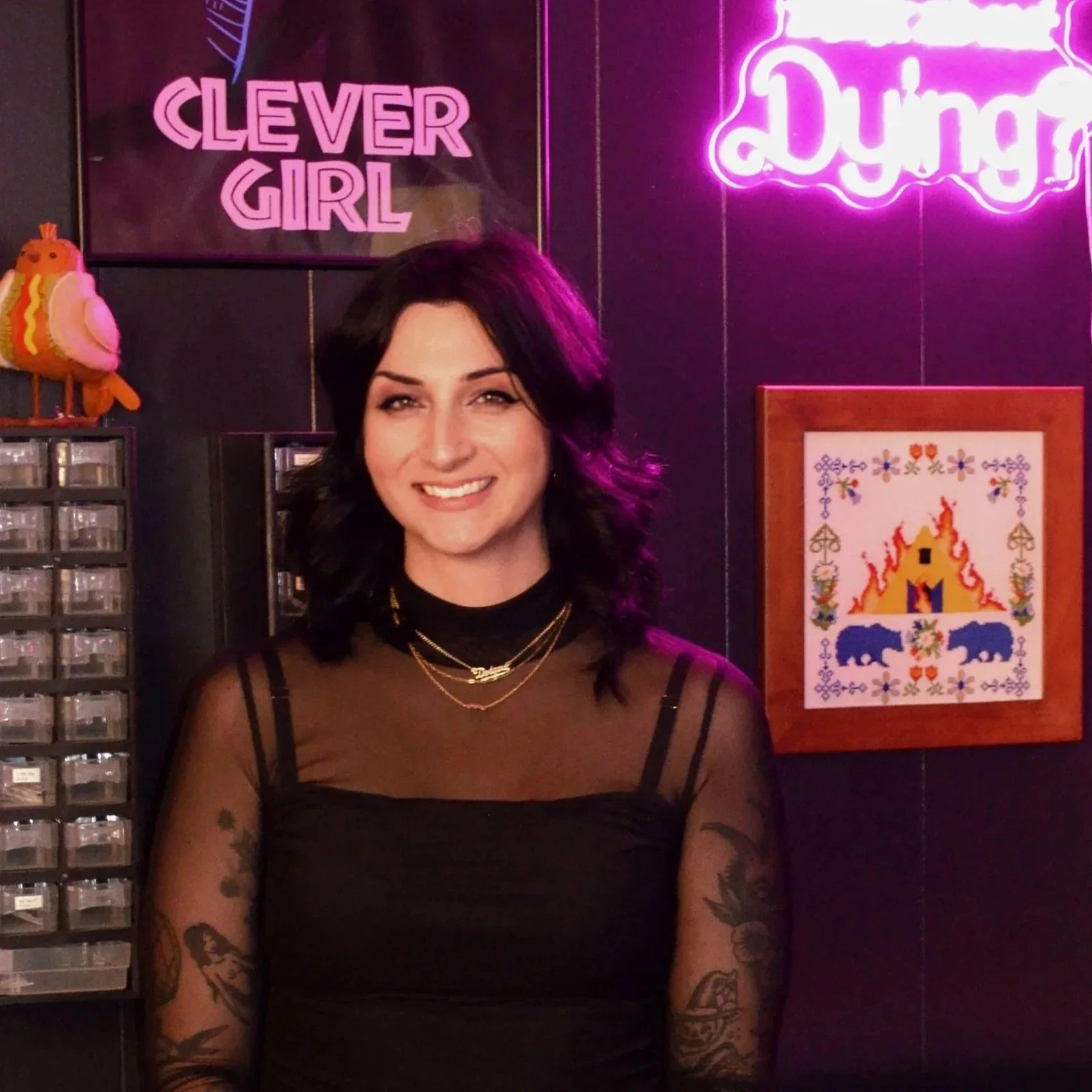 A woman with black hair and tattoos on her arms smiling in front of a neon sign that reads 'Clever Girl' and another sign that says 'Dying.' The background includes a framed embroidery piece with bear and fire motifs.