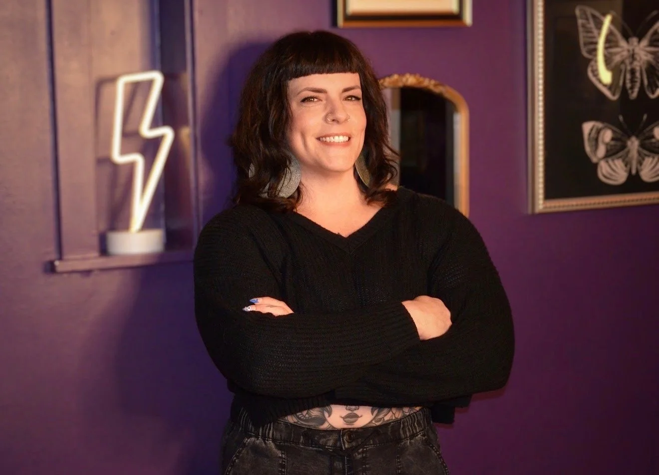 A woman with dark wavy hair and a nose piercing, smiling with arms crossed, standing in front of a purple wall with framed butterfly artwork and a neon lightning bolt sign.