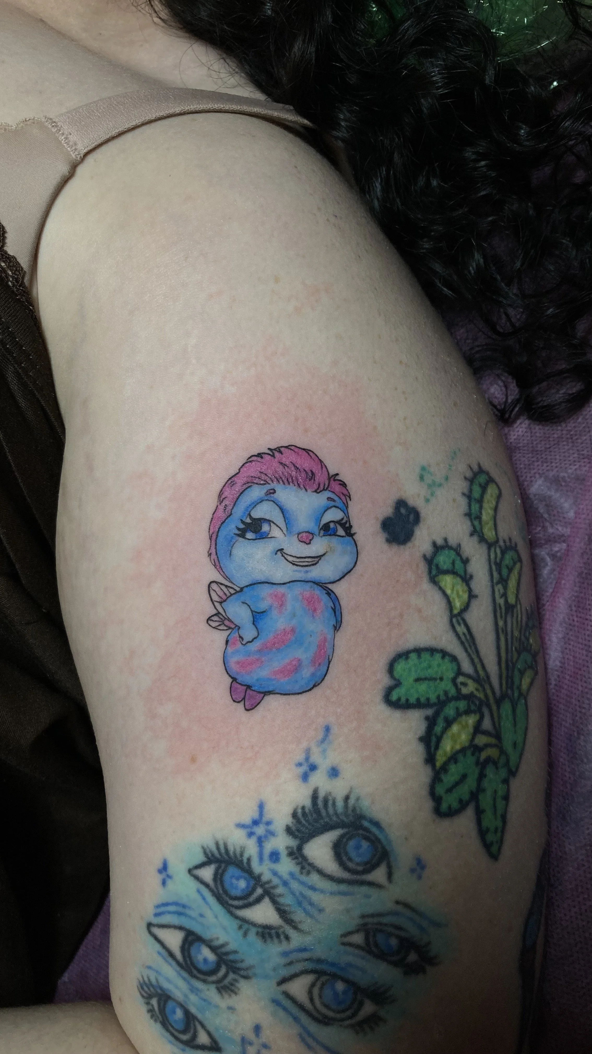 Tattoo of a cartoonish character with pink hair, blue fur, and a mischievous smile, next to a green plant with multiple leaves and buds, and a cluster of blue eyes with eyelashes below.