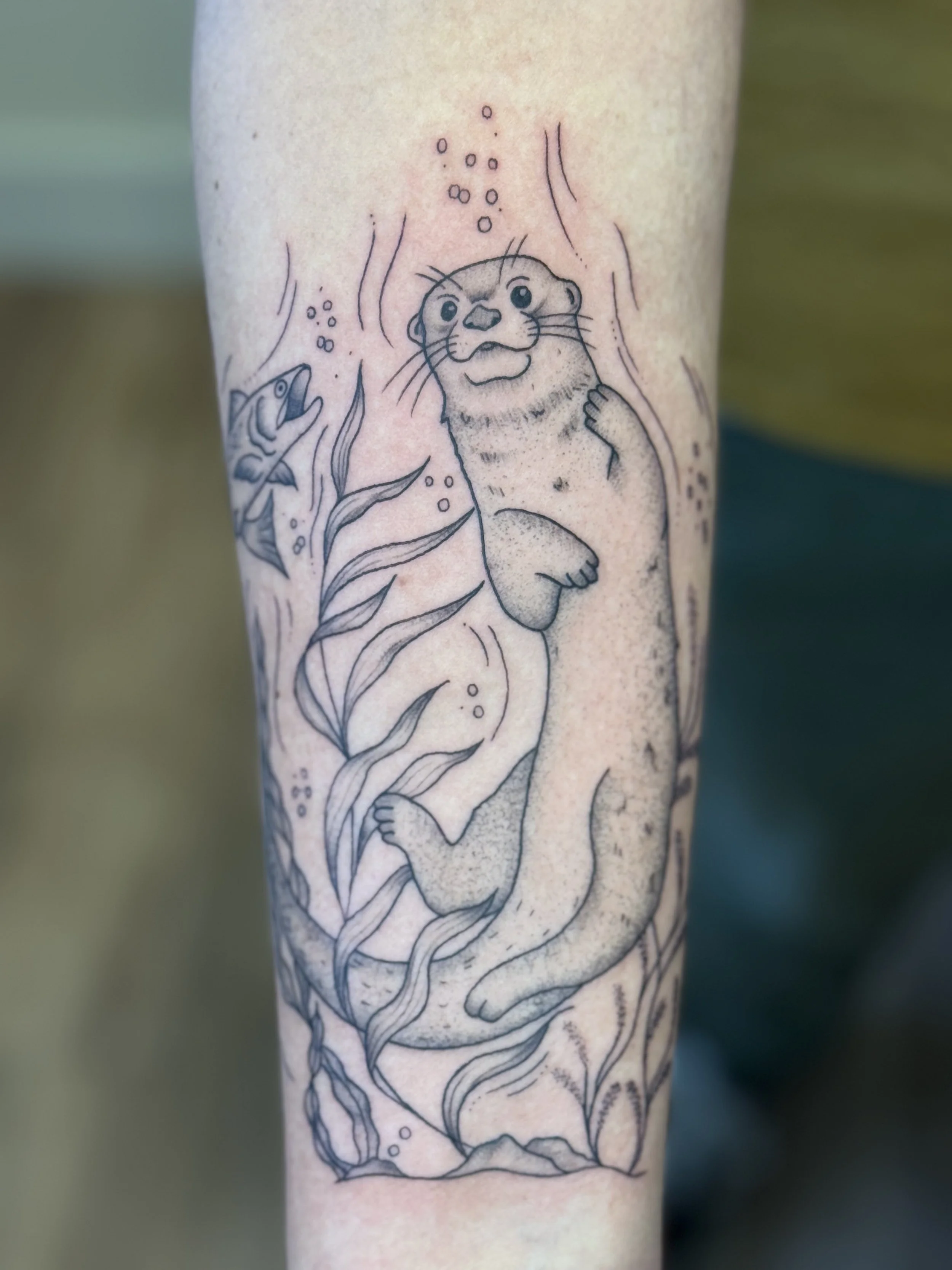 Tattoo of an otter underwater, surrounded by seaweed and a fish, in black ink outline style.