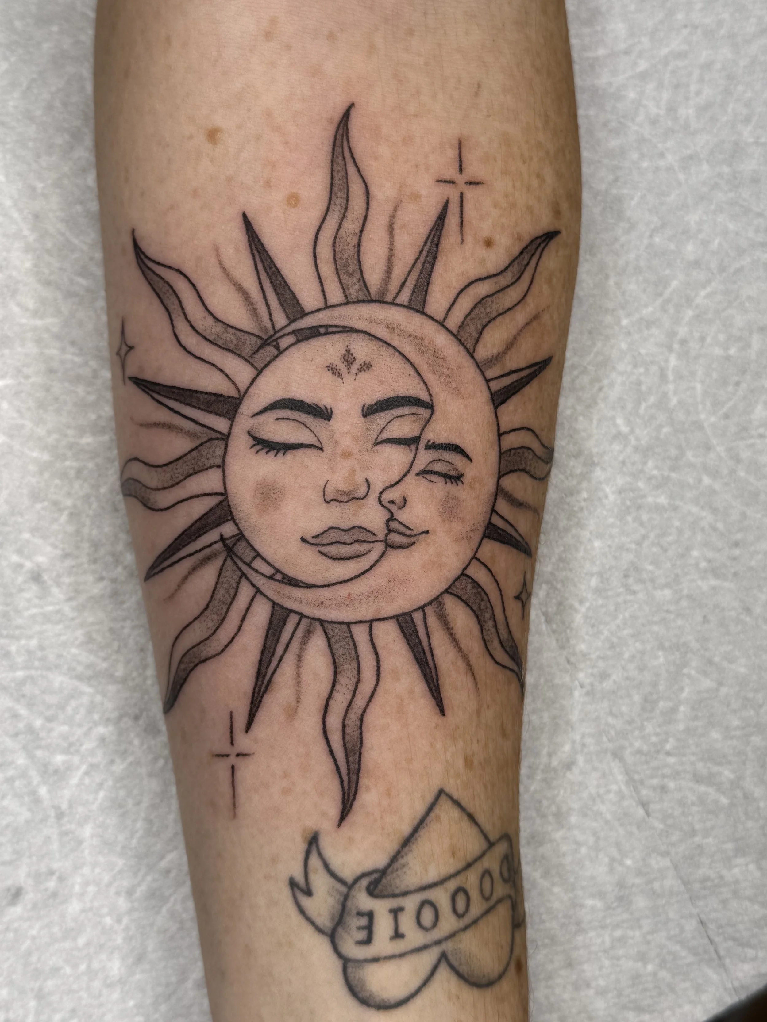 Tattoo of a sun with a smiling face, with a moon and a woman’s face with closed eyes inside the sun, and a banner with the word 'LOVE' below.