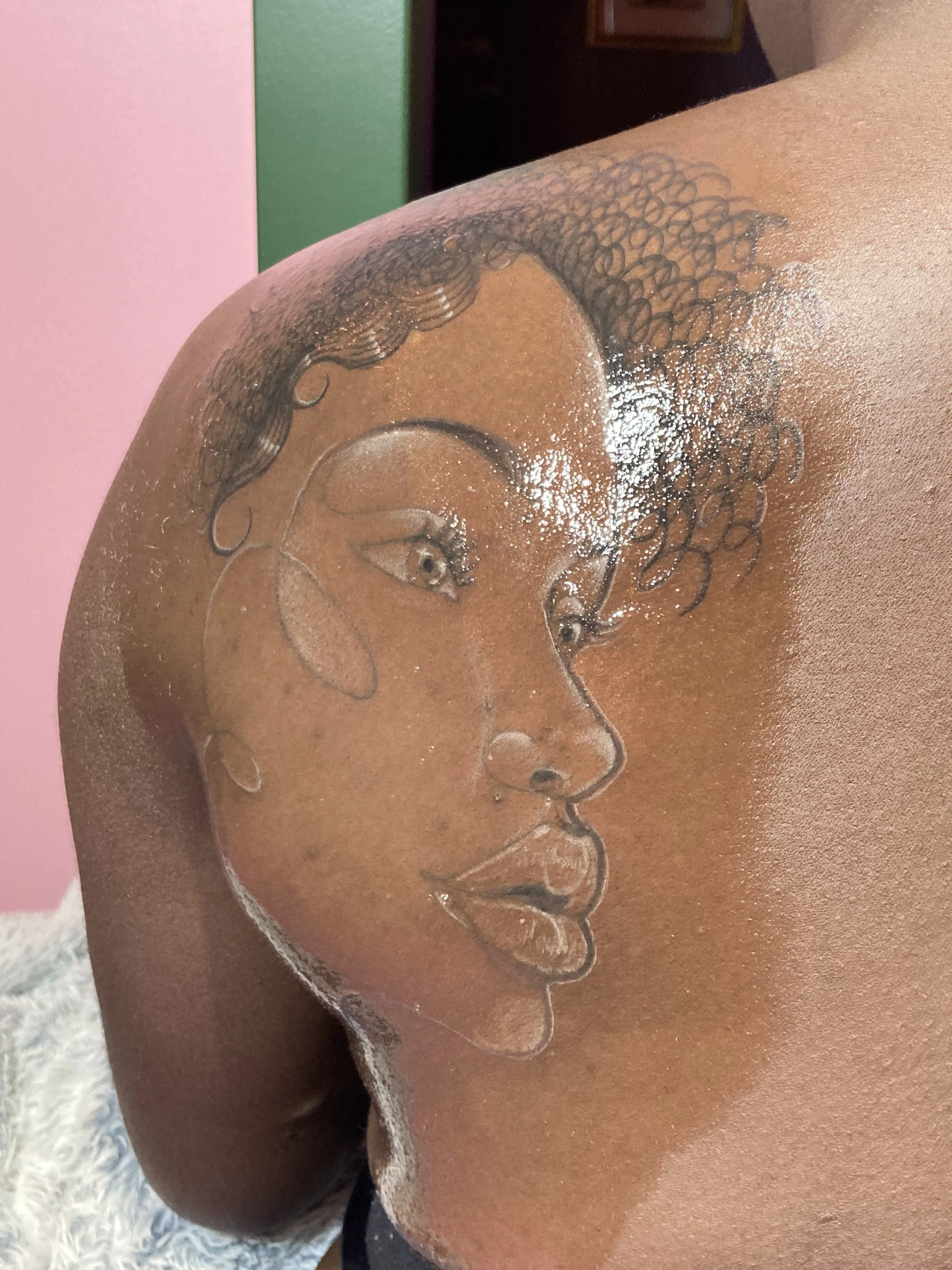 Tattoo of a woman's face on a person's shoulder, with curly hair, earrings, and expressive features.