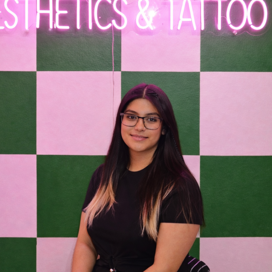 A young woman with long dark hair with blonde tips, wearing glasses and a black shirt, standing in front of a pink and green checkered wall with a neon pink sign that reads 'ESTHETICS & TATTOO'.