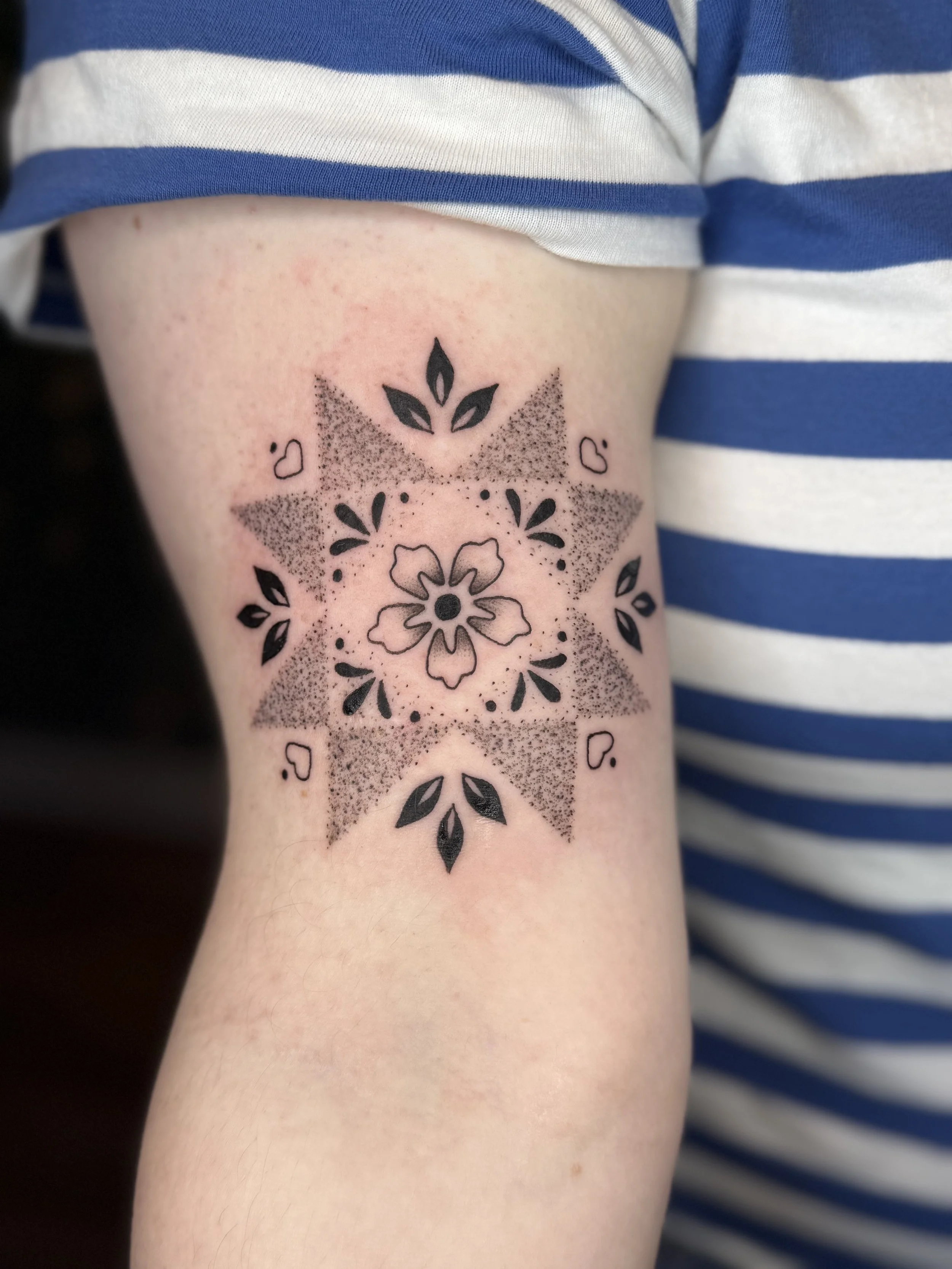 A geometric black ink tattoo on a person's upper arm, featuring a central flower surrounded by symmetrical patterns, stamped-style shapes, and leaf motifs, with shaded dot work creating depth.