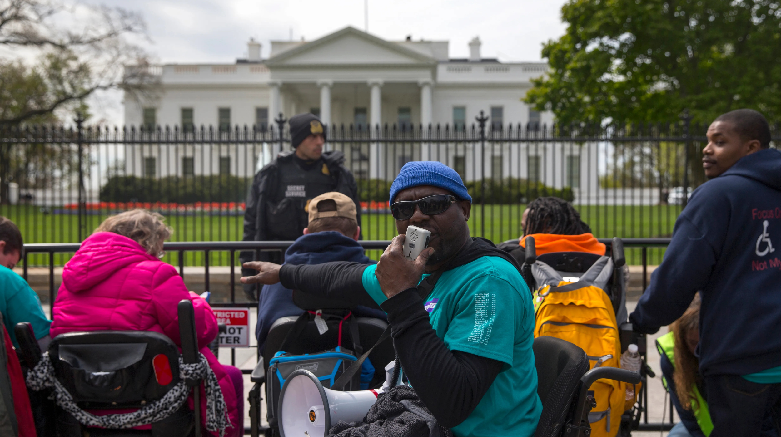 Protesters for disability rights sit along a barricade fence at the White House. Several are in wheelchairs, with oe protester facing the camera and speaking into a megaphone. A police officer stands in the background.