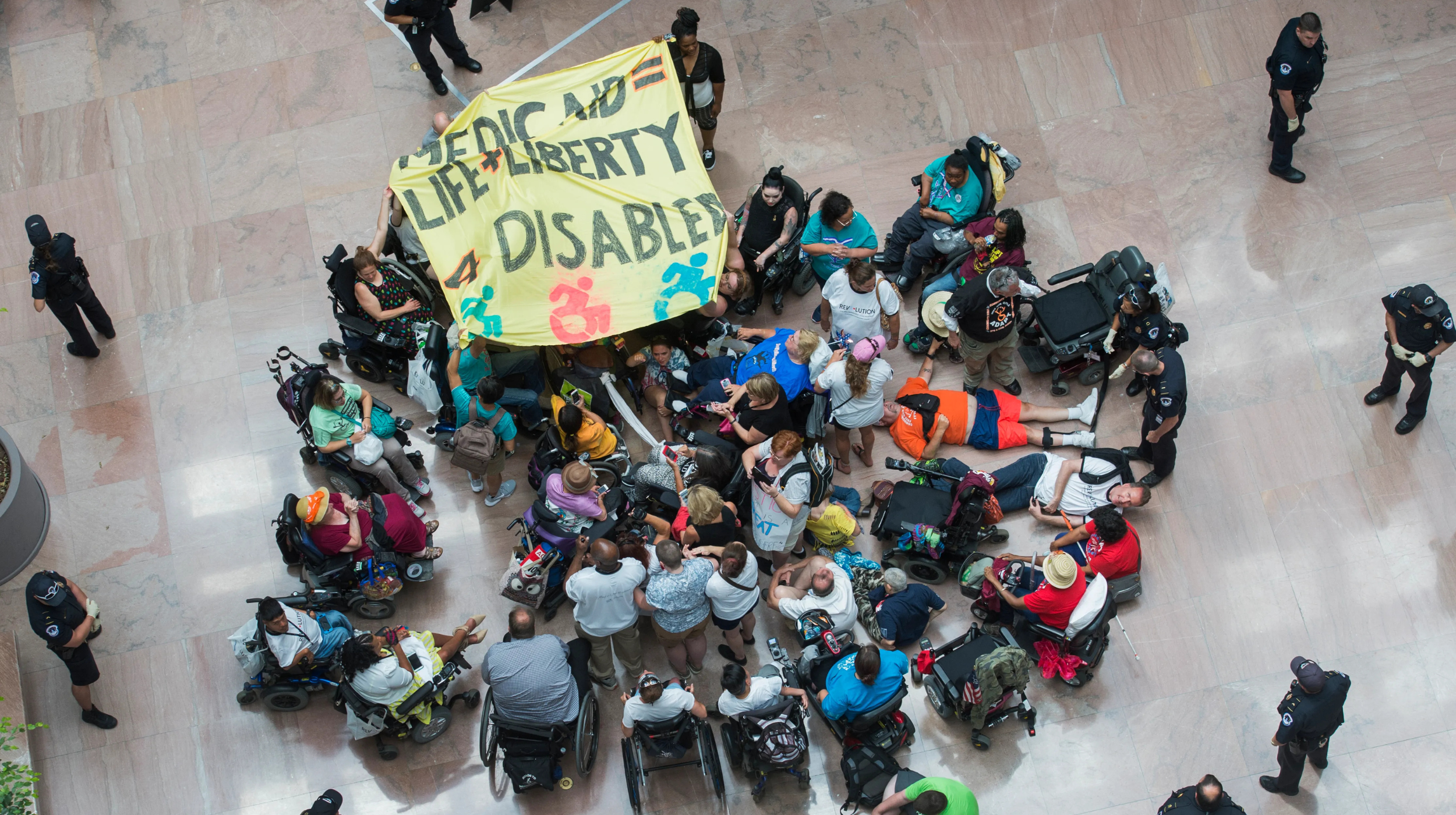 Many people gather in a public building during a protest. The group is arranged in a circle, some sitting in wheelchairs, others sitting or lying on the floor. They hold a large sign that reads, "Medicaid equals life and liberty for disabled."