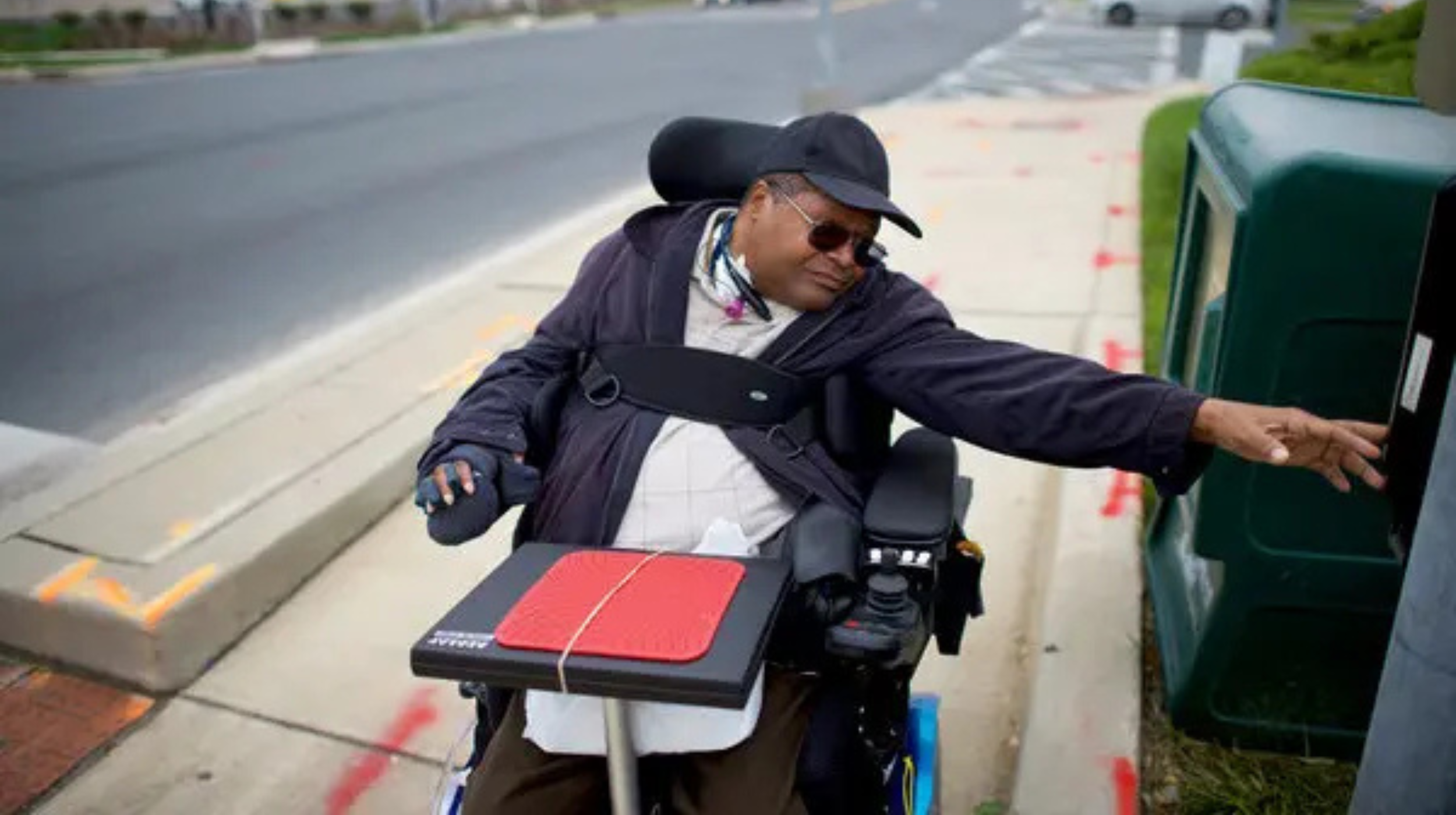 A person dressed in. a black jacket, hat, and sunglasses reaches for a newspaper box from their wheelchair on a sidewalk.