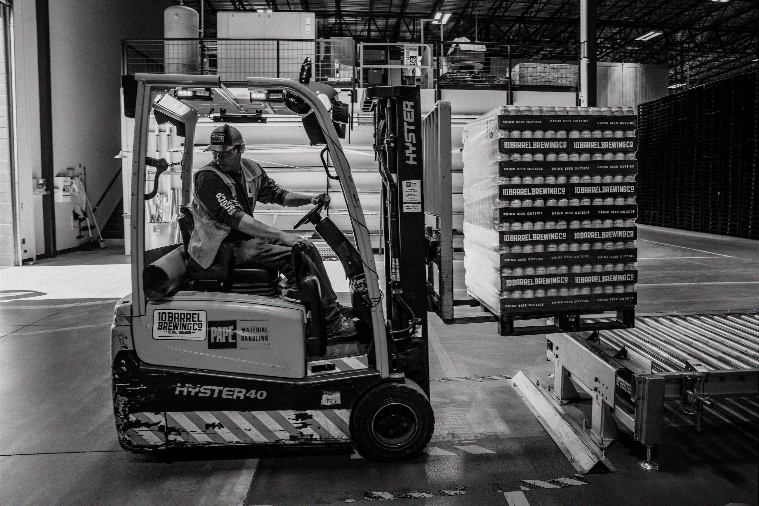 A person operates a forklift, removing a loaded palette from a conveyor belt.