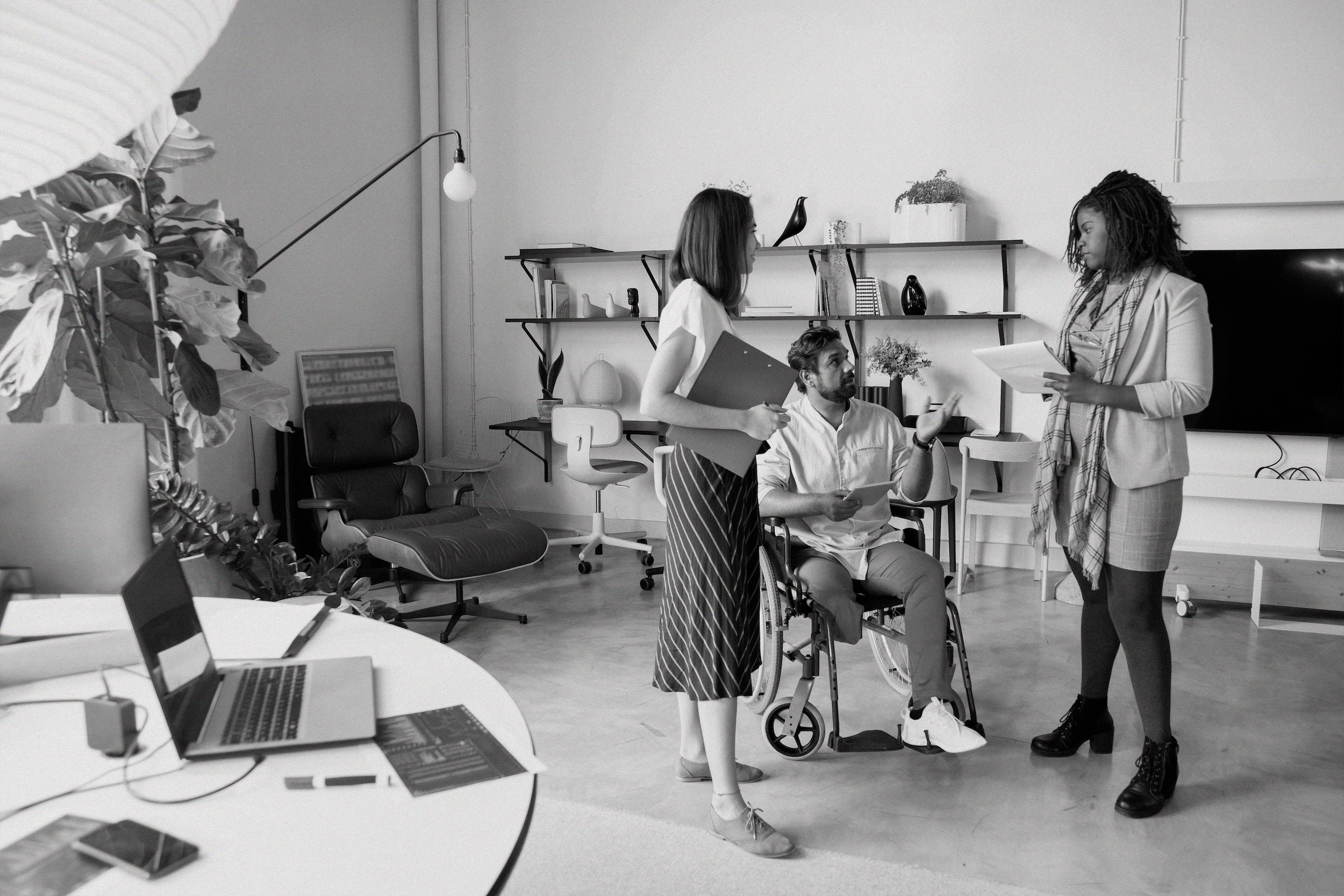 Three people interact in an office - two are standing and one is using a wheelchair.