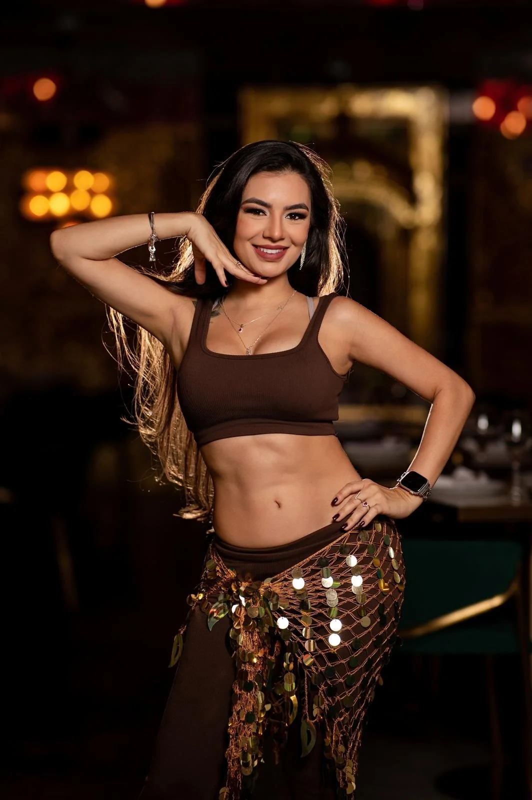 A woman smiling and posing confidently in a dimly lit venue, wearing a brown crop top and a decorative skirt with hanging reflective discs, gold jewelry, and an Apple Watch.