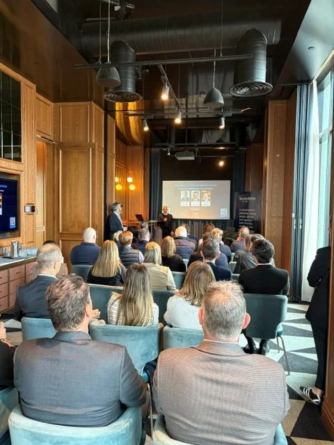 Business conference or presentation with attendees seated facing a speaker and presentation screen in a modern, wood-paneled room with large windows.