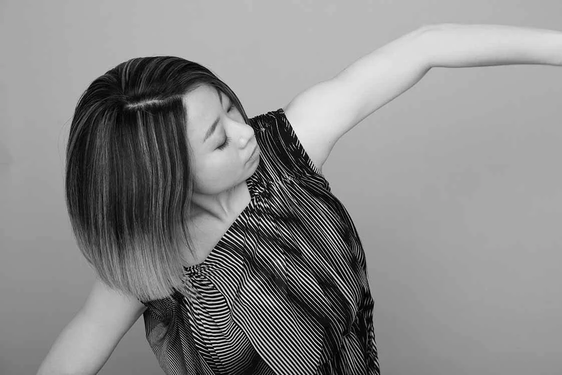 グレーの背景で黒と白のストライプ柄のドレスを着た女性が左側を向き、腕を伸ばしている姿を写した黒と白の写真