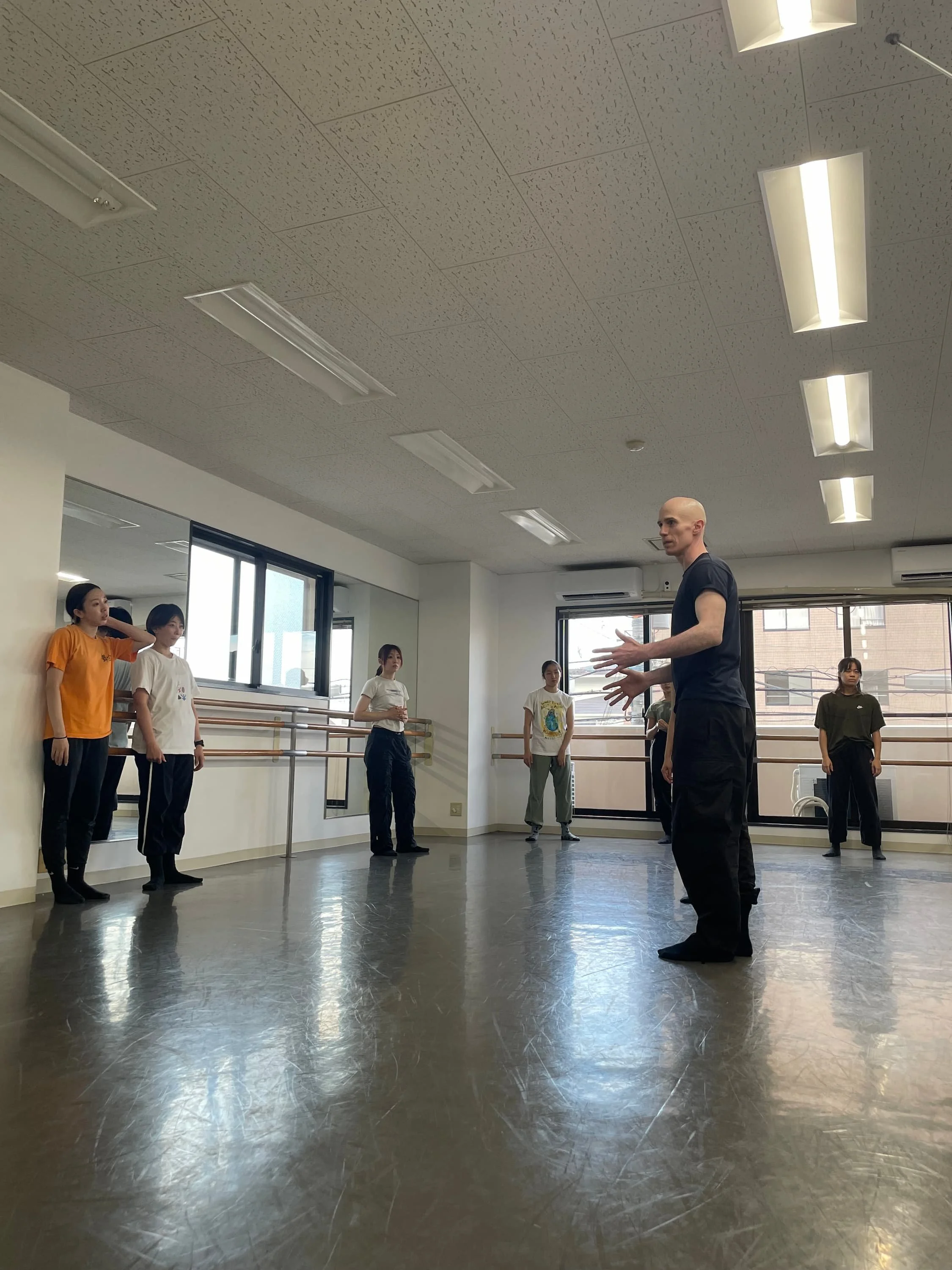 ダンスのレッスンをしている男女のグループと、リーダーらしき男性が指導している室内風景。壁に鏡と手すりがあるスタジオ。