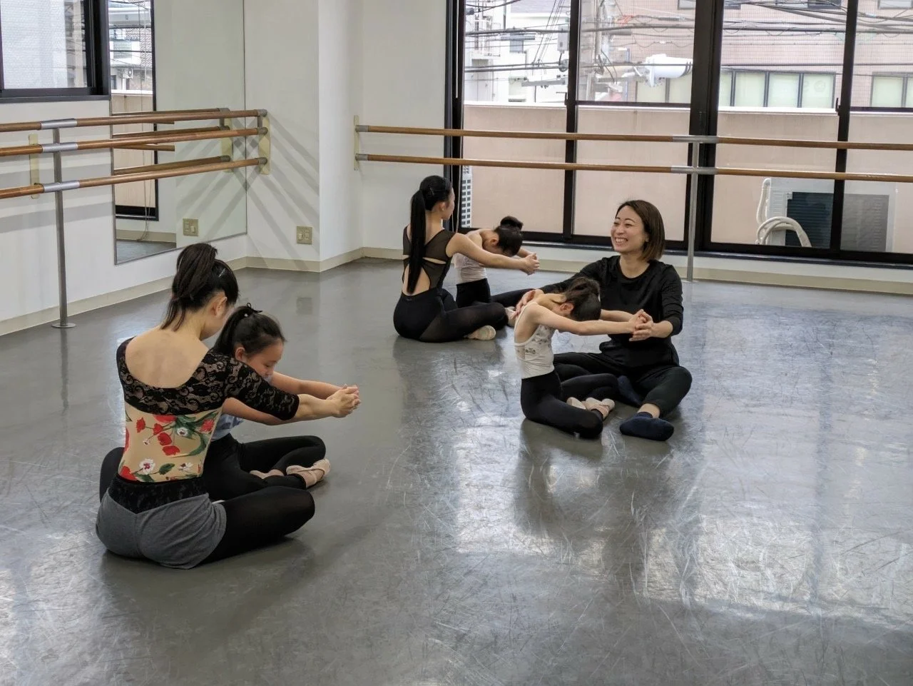 バレエレッスンをしている大人と子供のグループ。子供たちは先生の指導のもとで座って前屈をしている。