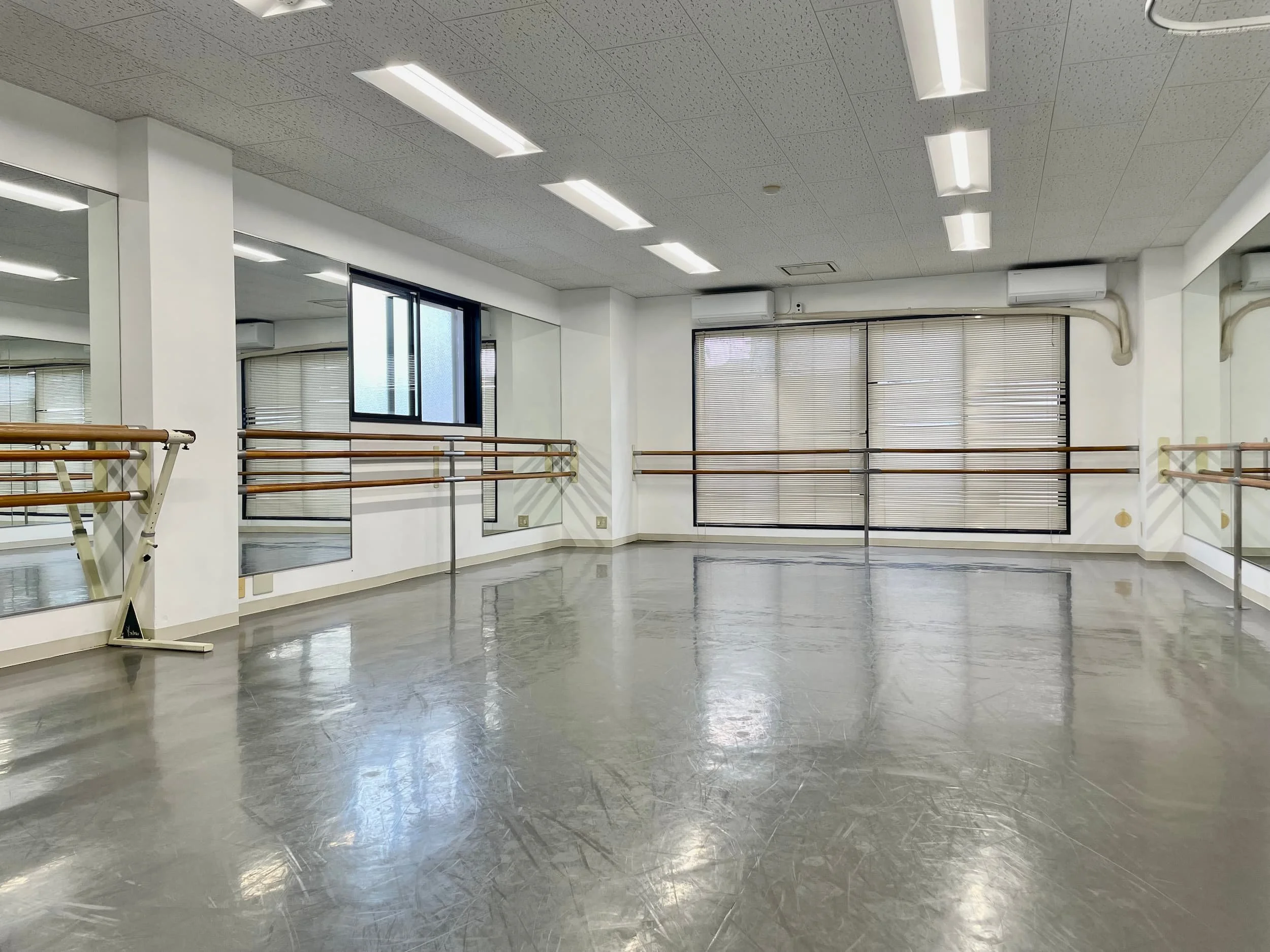 空のダンススタジオまたはリハーサルスタジオ。鏡とバレエの棒が壁に設置されている。大きな窓と明るい照明があり、広く、清潔な空間。