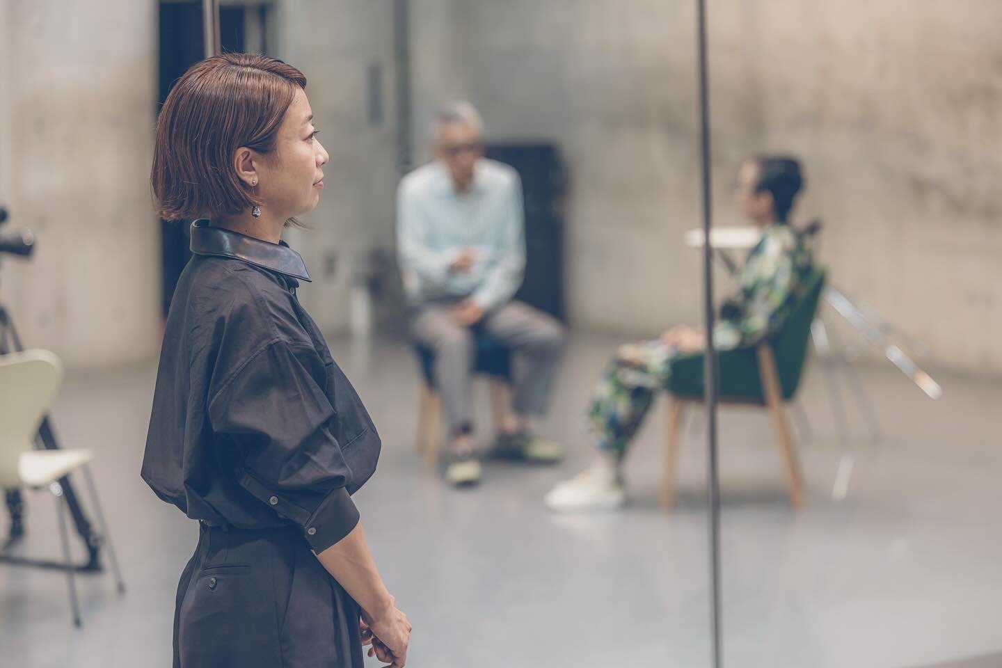 若い女性が黒い服を着て、鏡の前で立っている写真。背景には椅子に座る2人の人がいる。