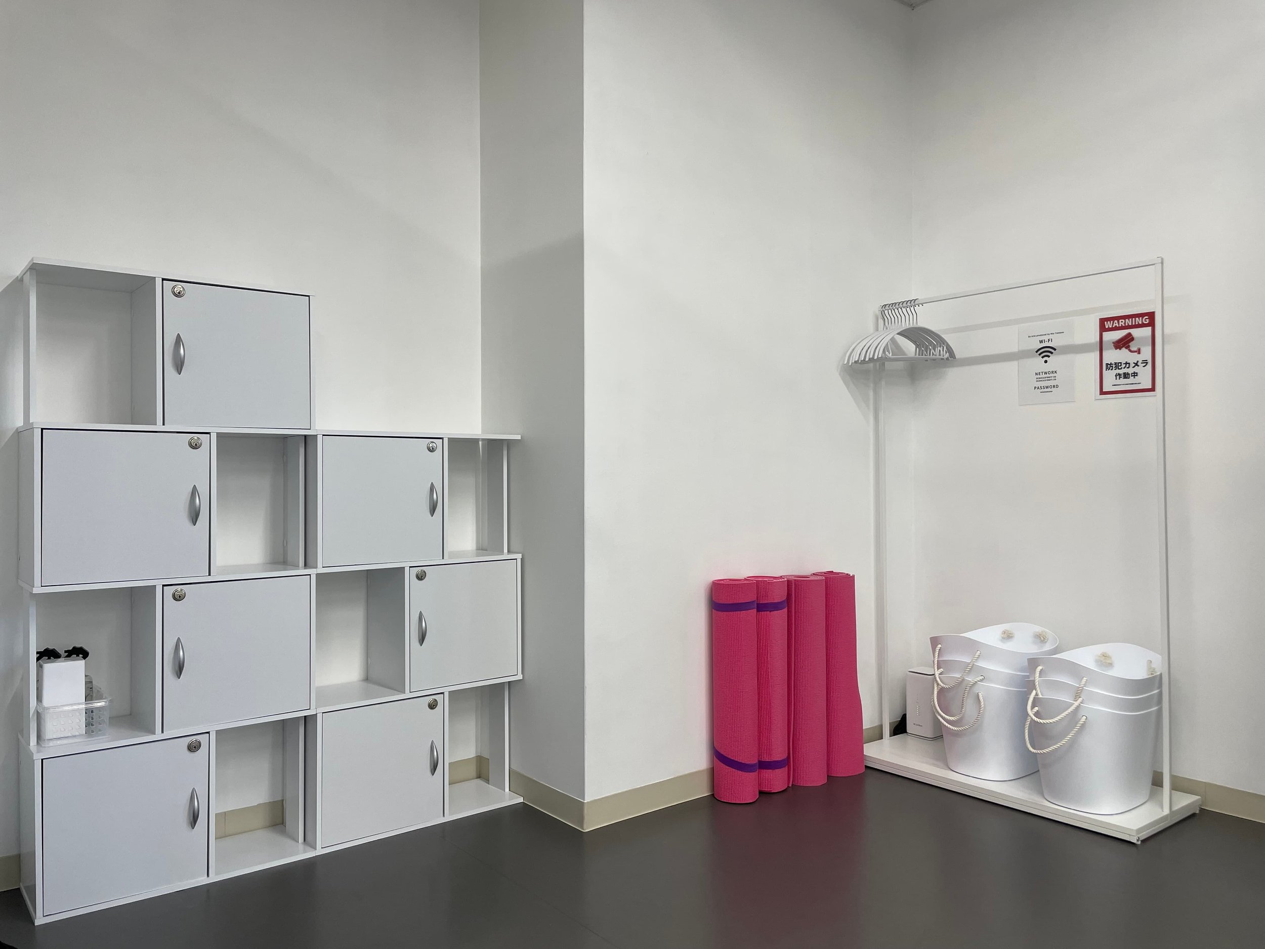 白いロッカー棚、ピンクのヨガマット、白いバスケットとバッグ、掲示板に防犯カメラ作動中の注意書きとWi-Fi情報の紙があるフィットネスジムの更衣室の一角です。