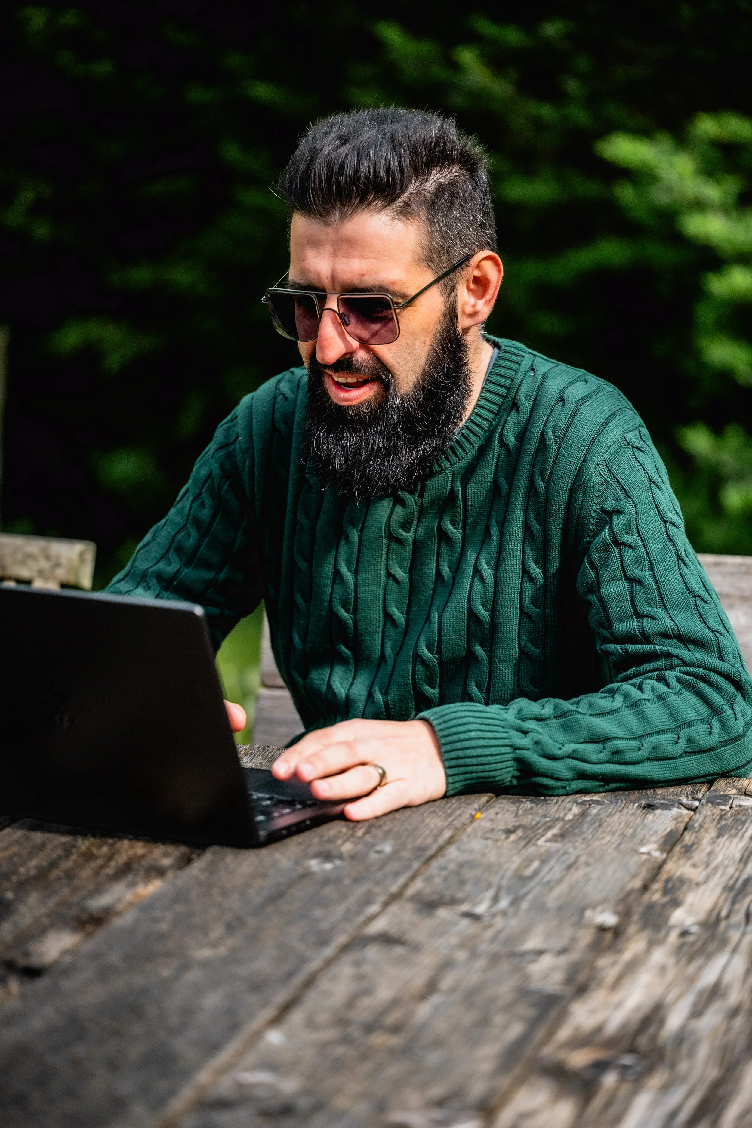 Man with dark hair and beard wearing sunglasses and a green cable-knit sweater, working on a laptop at an outdoor wooden table in a natural setting.