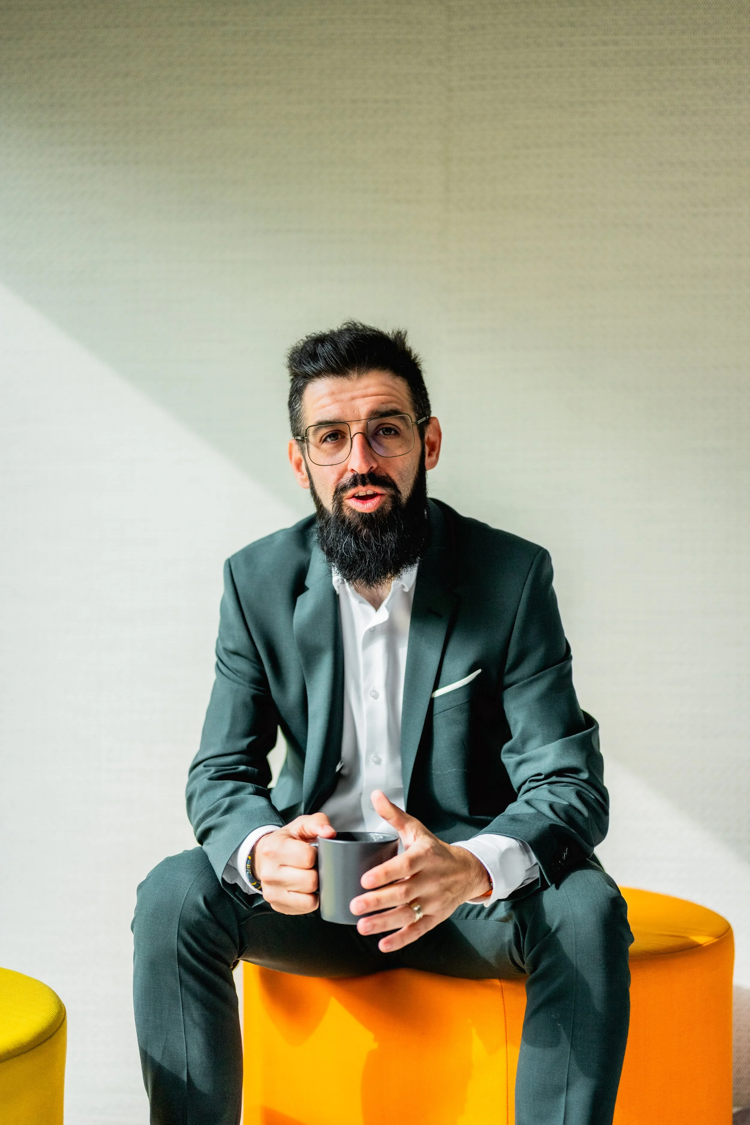 A man with dark hair, glasses, and a beard wearing a dark suit and white shirt, sitting on an orange chair, holding a black mug, in a modern office setting with colorful seating around him.