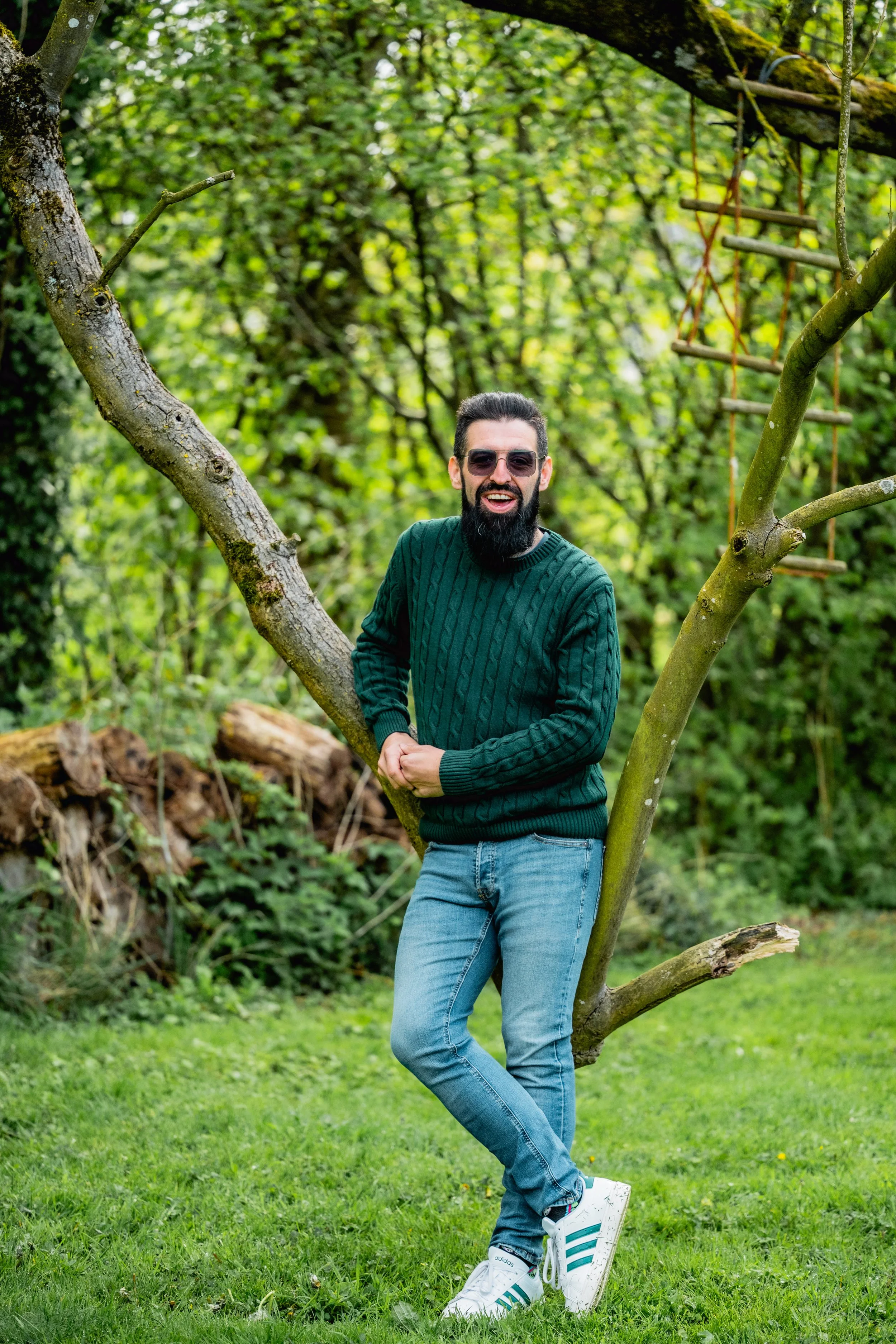 A man with a beard and sunglasses standing outdoors on grass, leaning against a tree with branches. He is wearing a green sweater, blue jeans, and white Adidas sneakers with turquoise stripes, and is smiling.