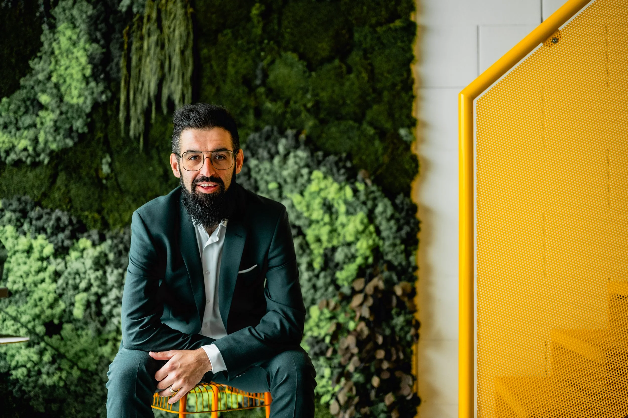 A man with black hair, glasses, and a beard, wearing a dark suit and white shirt, sitting on an orange metal chair indoors next to a green leafy vertical garden wall and yellow partition.