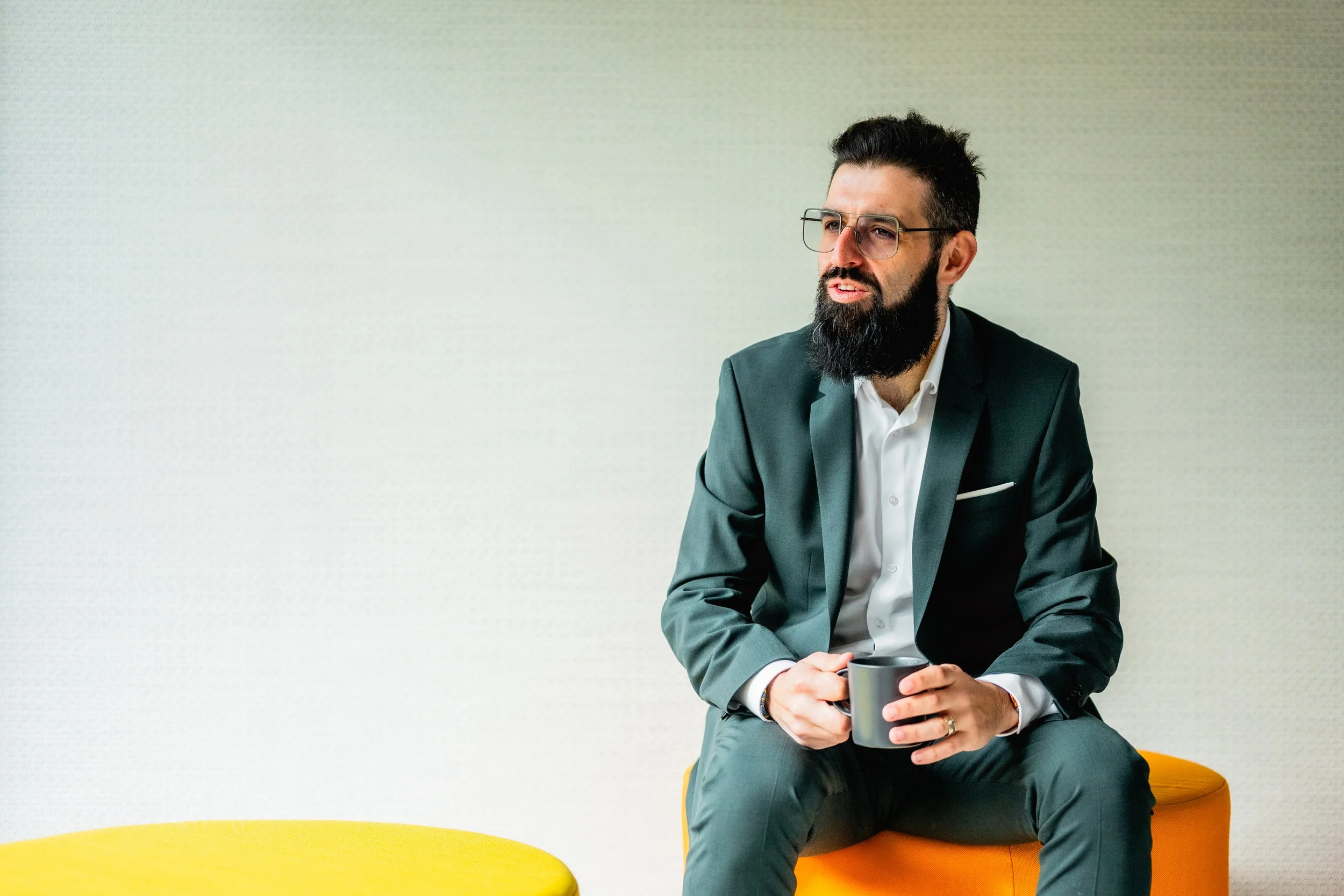Man with dark hair, glasses, and a beard wearing a dark suit and white shirt, sitting on an orange cushioned seat, holding a coffee mug, against a plain light-colored wall.