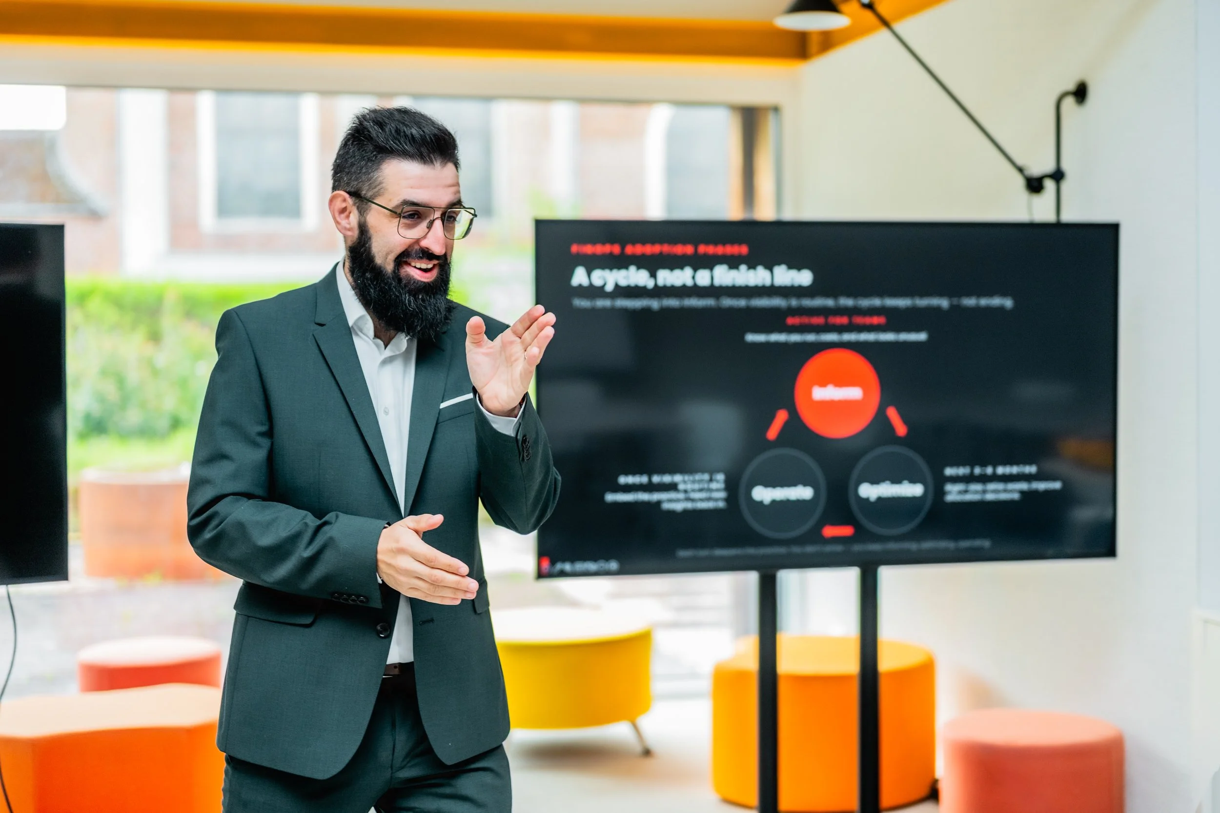 A man with dark hair, glasses, and a beard wearing a gray suit and white shirt, standing in front of a large screen in a modern office or conference space with large windows and colorful seating.