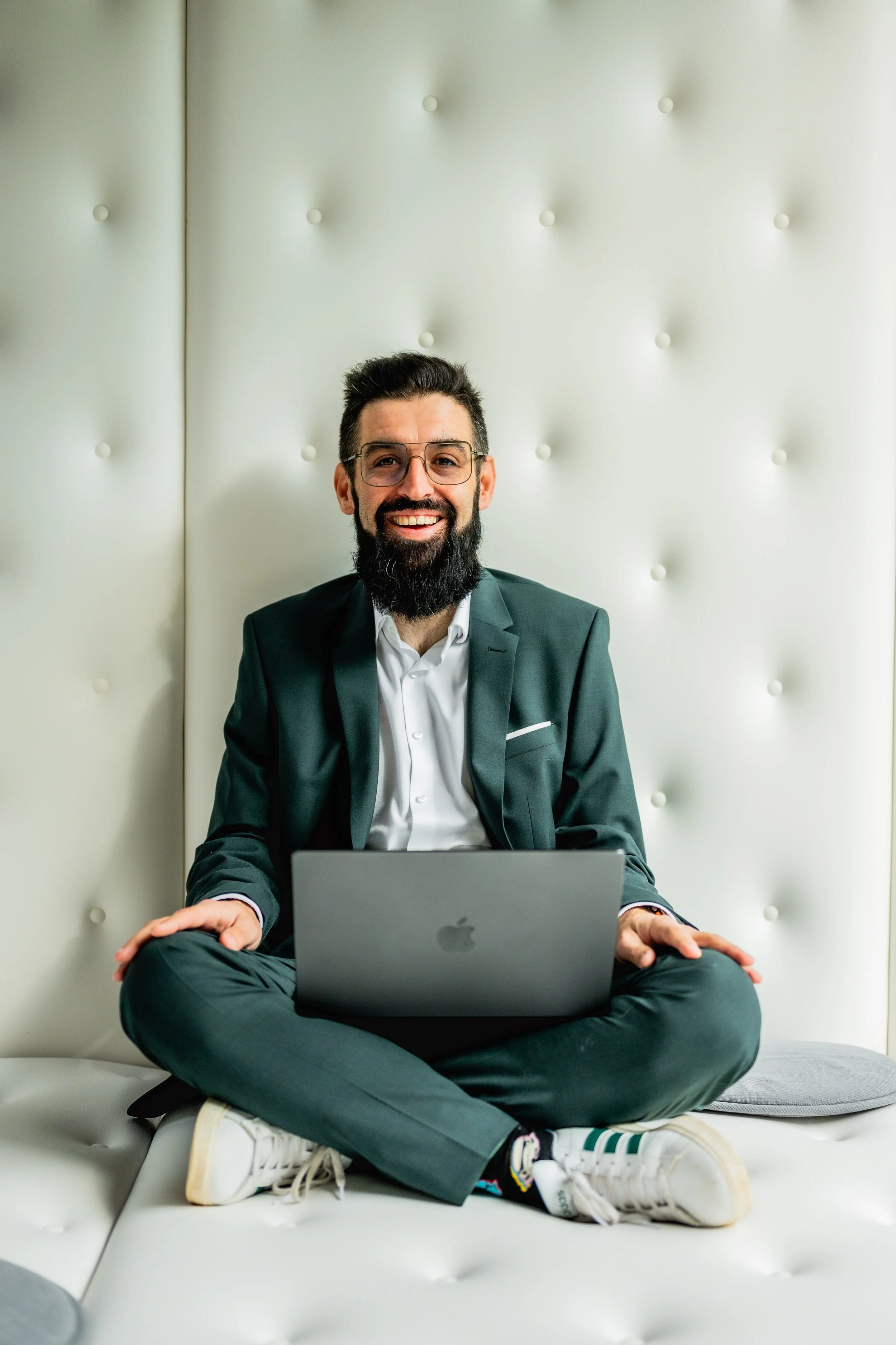 A man with dark hair, a beard, and glasses, dressed in a suit, sitting cross-legged on a white sofa and working on a gray laptop.