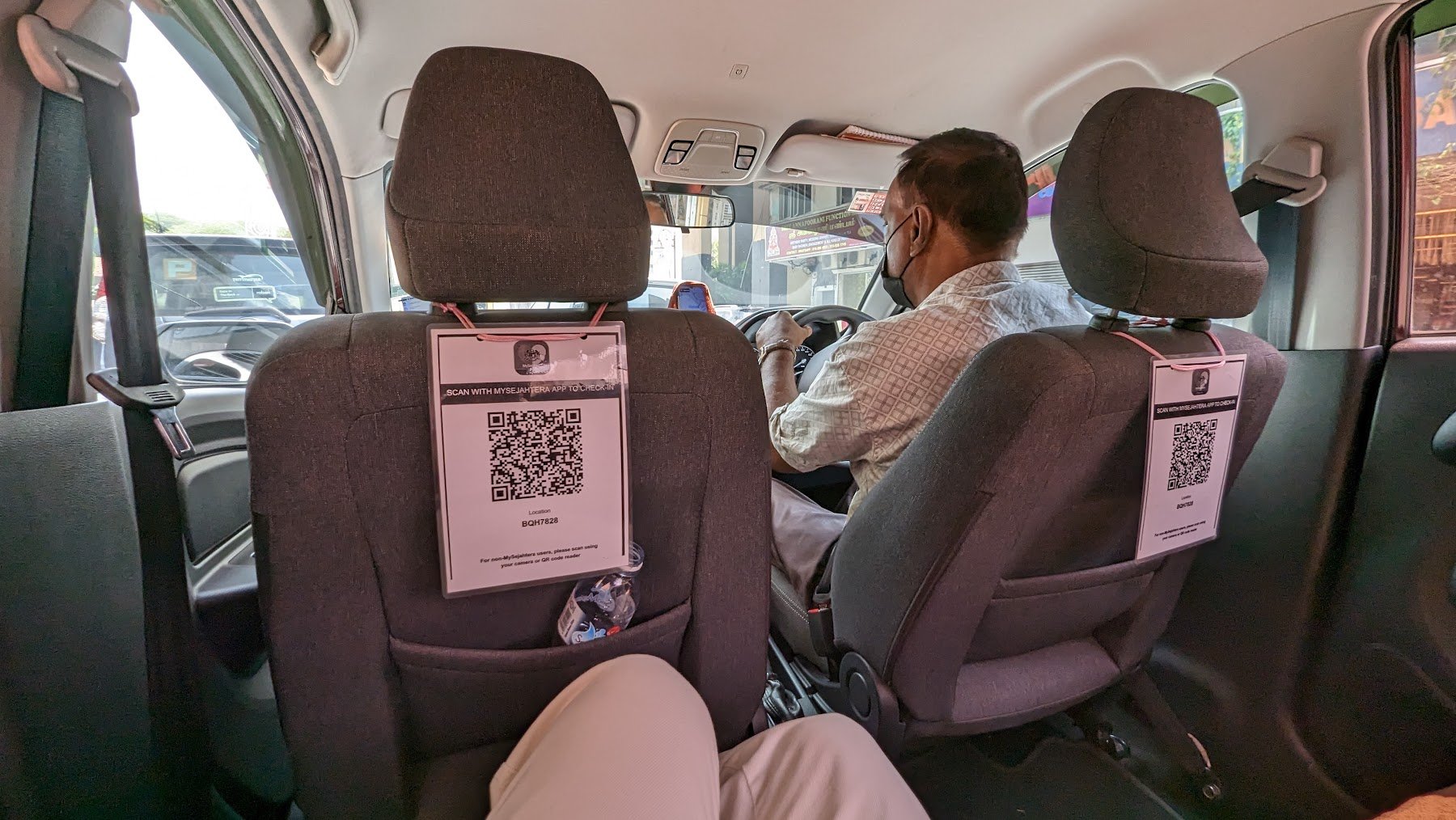 The view from the backseat of a rideshare car, with two grey seats in front, a driver wearing a mask, QR codes hanging from the headrests, and a city scene outside