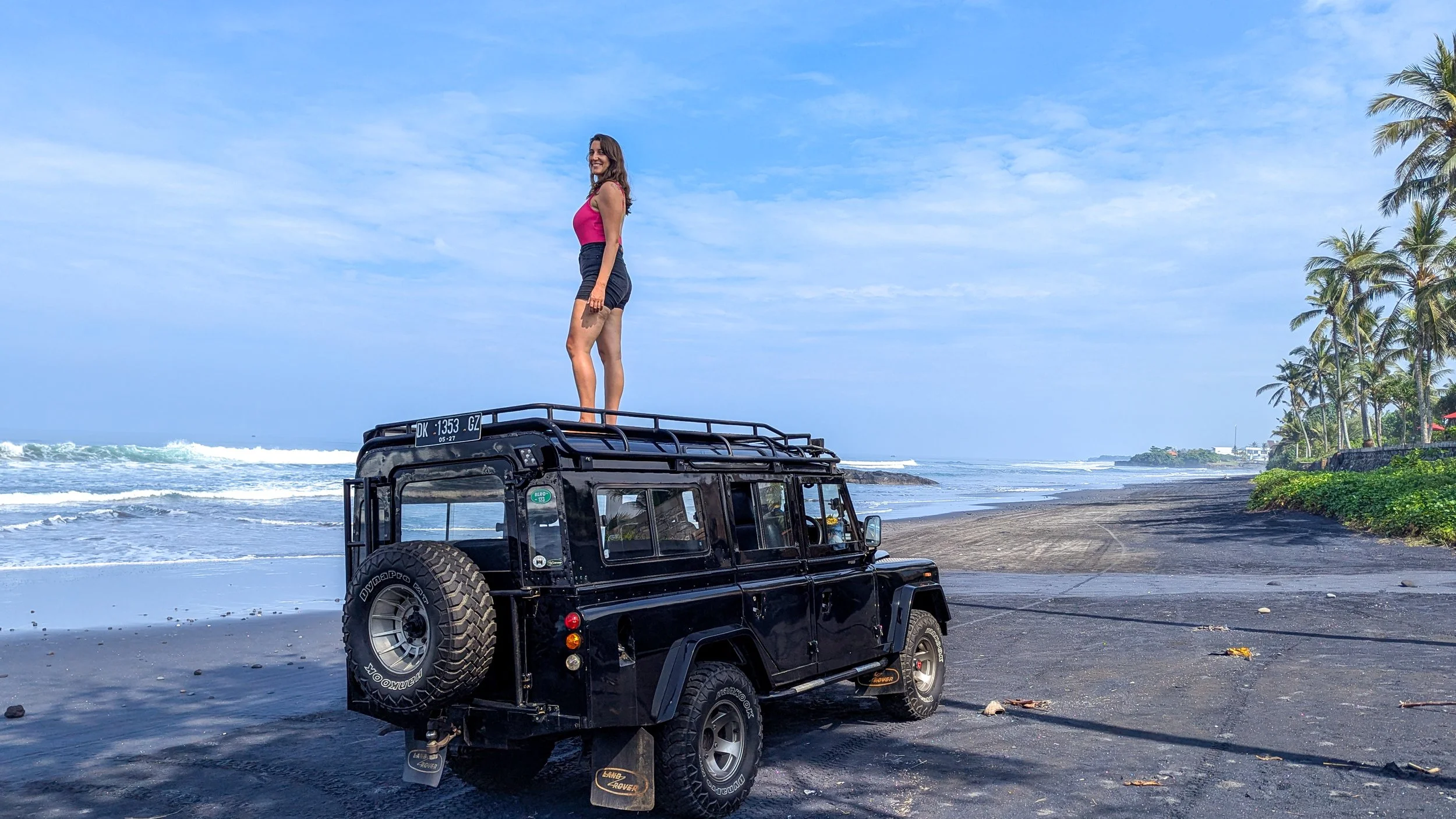 a woman stands on top of a black land rover on a black sand beach in Tabanan Bali
